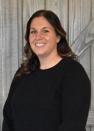 Woman smiling, wearing a black long-sleeved shirt, in front of a gray wood background.