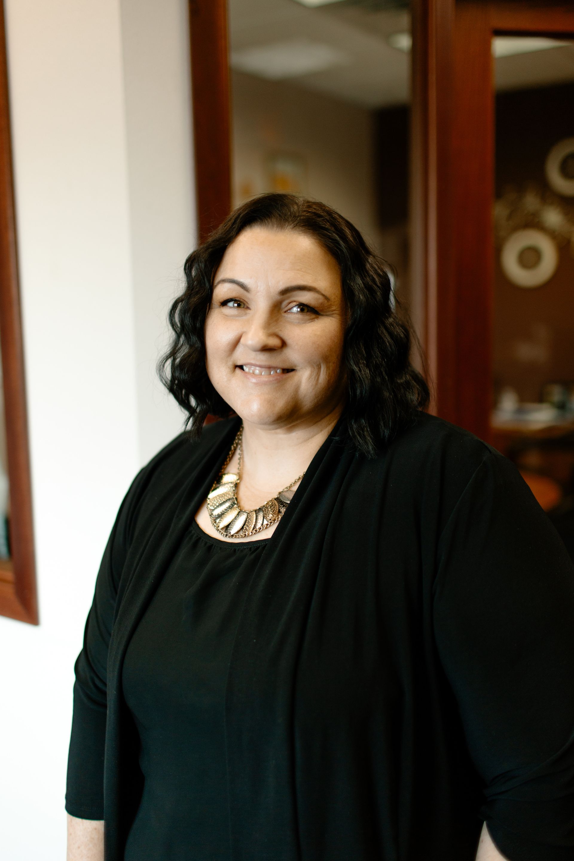 Woman in black cardigan and necklace smiles in an office setting.