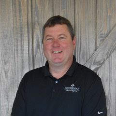 Man in a black polo shirt, smiling, standing in front of a wood background; shirt says