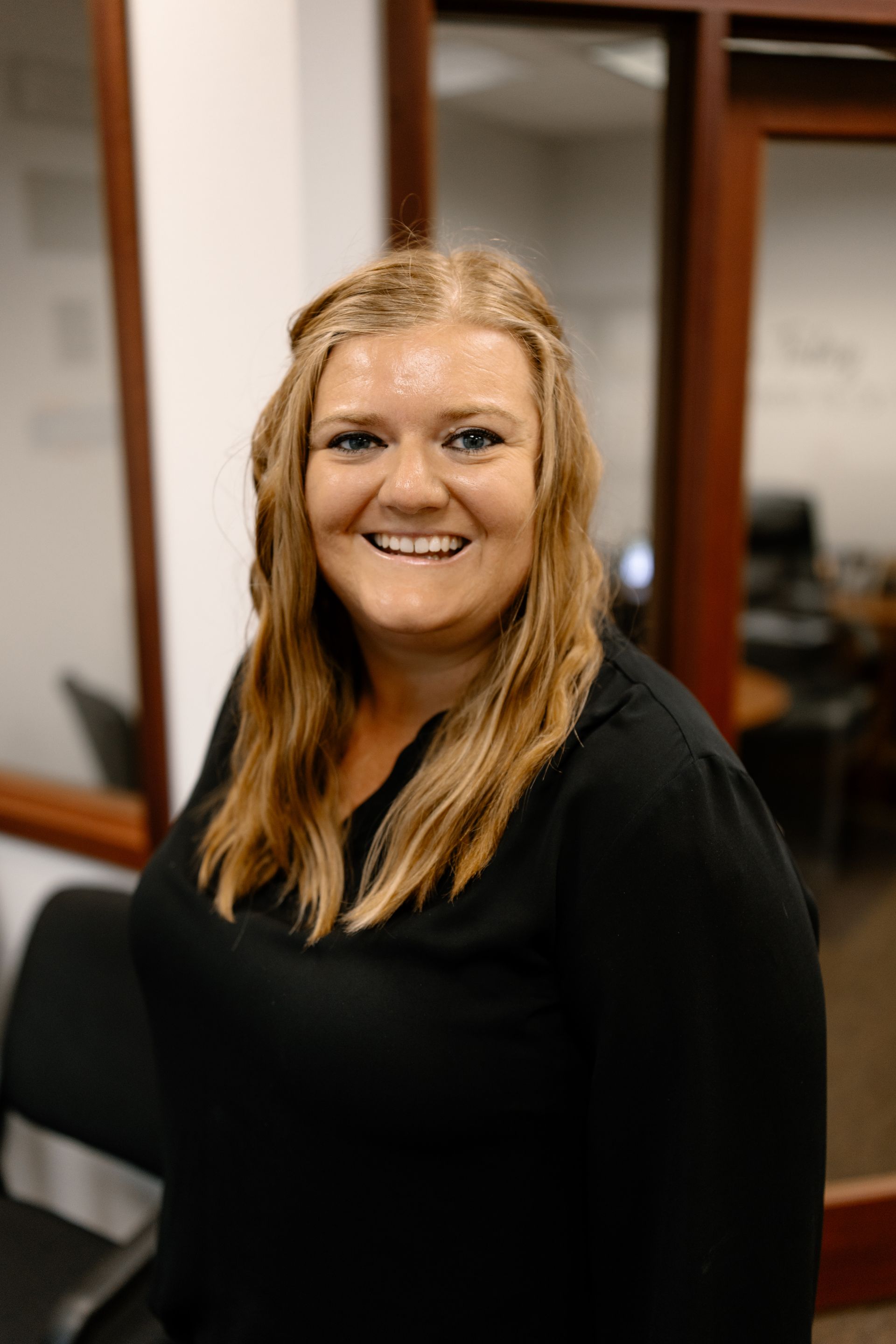 Woman with blonde hair, smiling, wearing a black top, in an office setting.