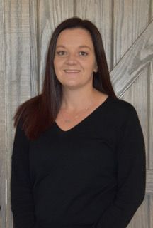 Woman with long brown hair, wearing a black v-neck shirt, smiling in front of a light wood backdrop.