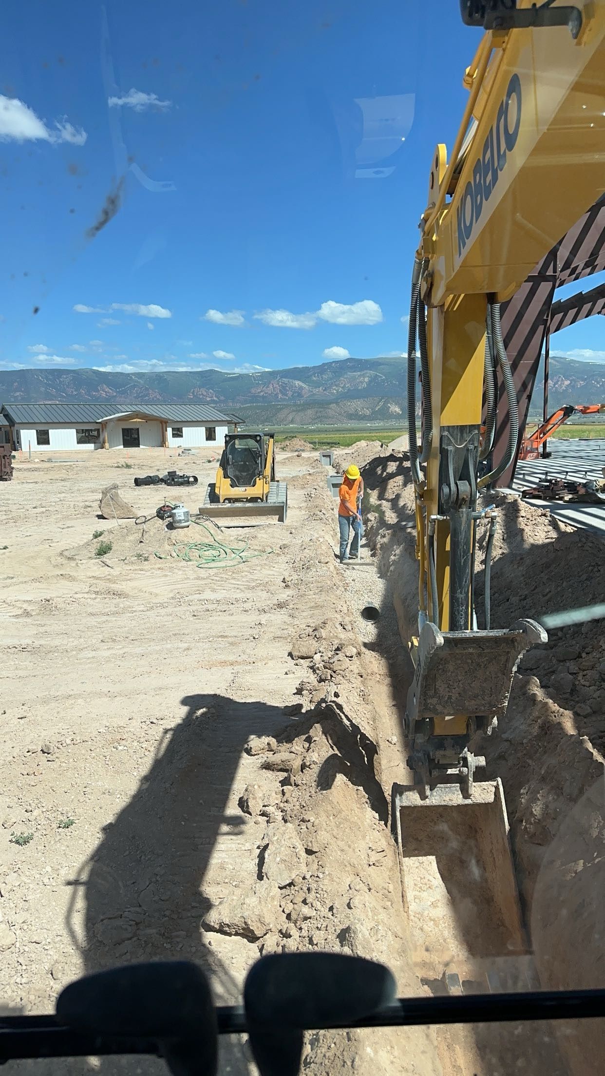 Construction site with a yellow excavator digging a trench. A worker in orange vest and hard hat stands nearby. Mountains in background.