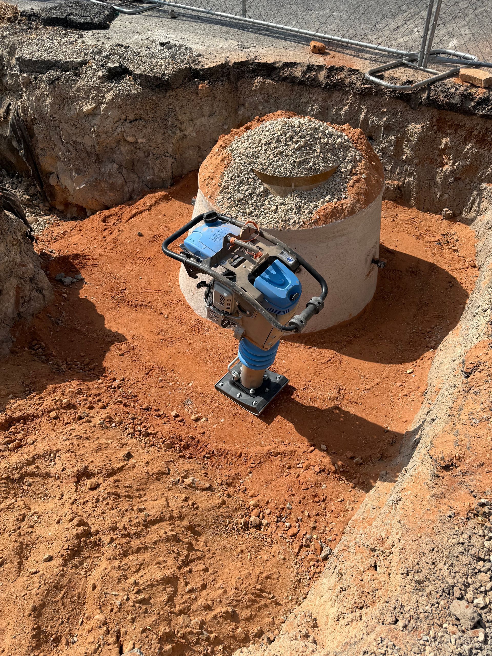 A construction site with a compactor tool on reddish soil, preparing the ground around a concrete structure filled with gravel.