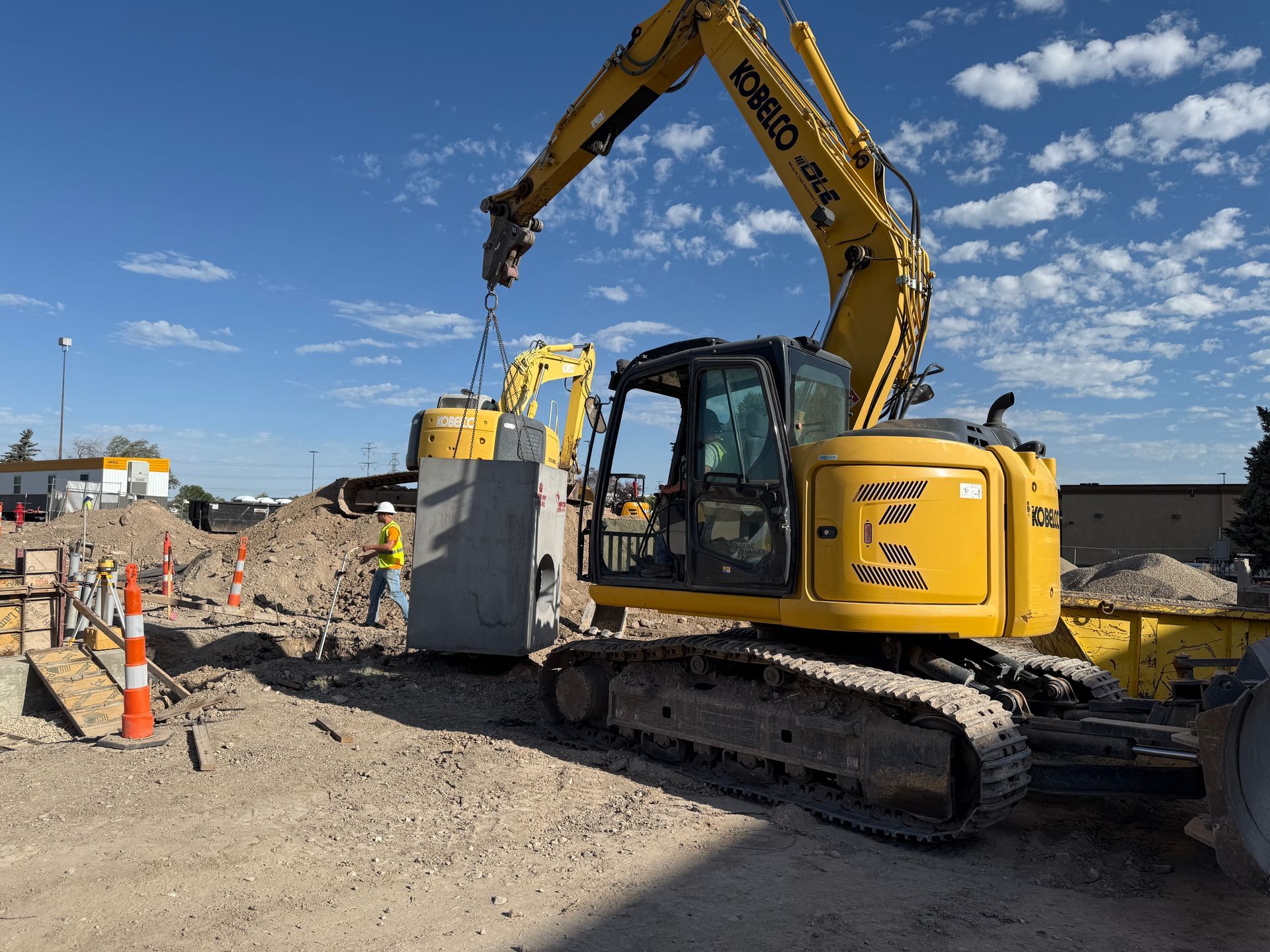 A yellow excavator lifts a large gray slab at a construction site on a sunny day. A worker stands nearby.