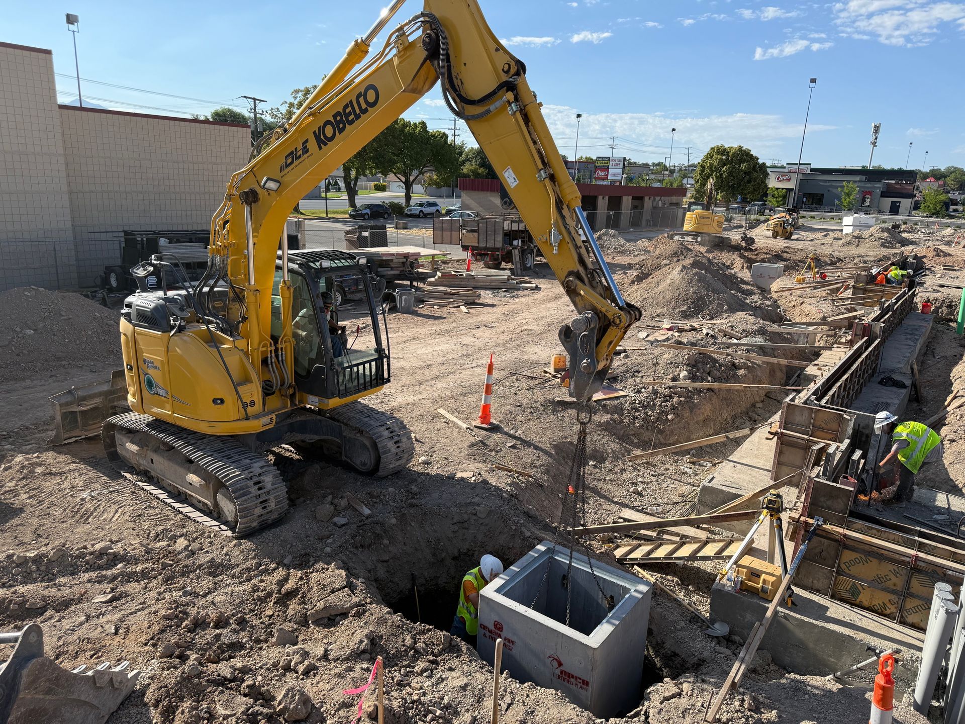 A yellow excavator digs at a construction site, with workers near a concrete structure in the hole. Sunny outdoor setting.