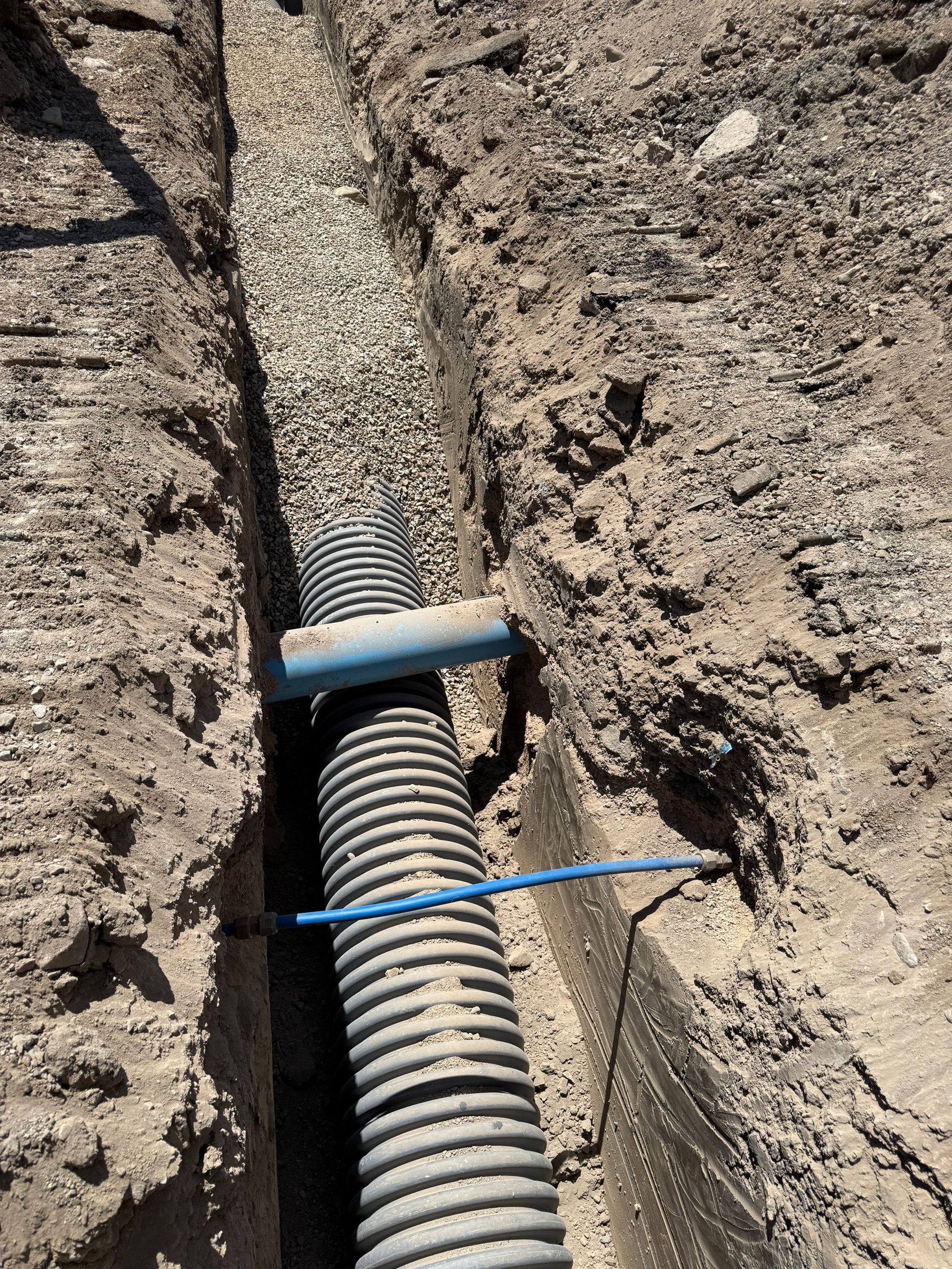 A narrow trench holding a corrugated black pipe connected to a blue pipe. The trench is filled with gravel.