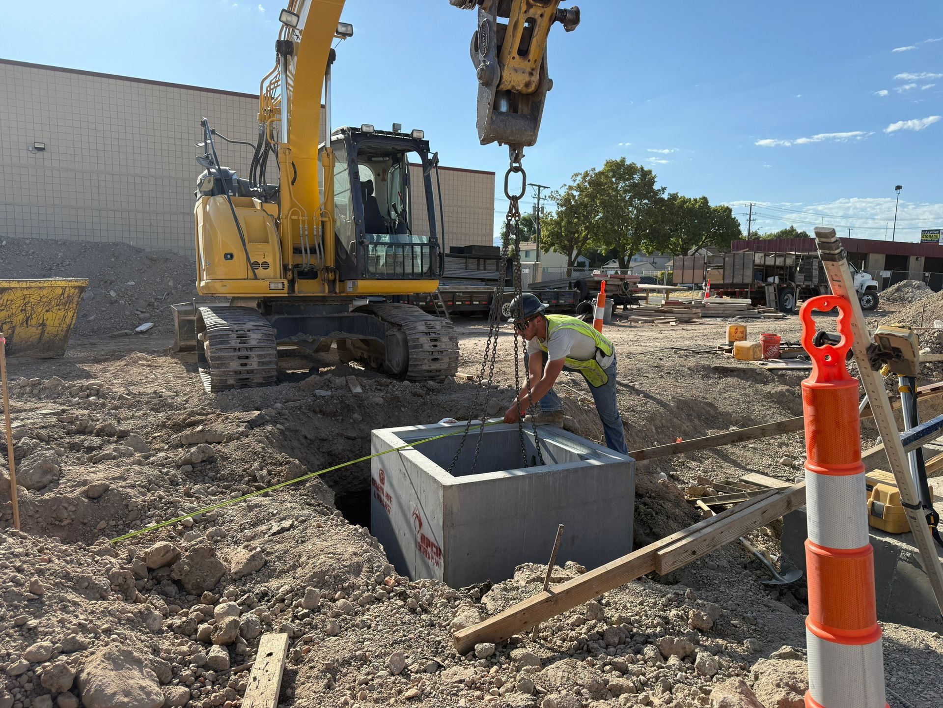 Construction worker positioning a concrete box in a dug pit with a yellow excavator overhead on a construction site under a blue sky.