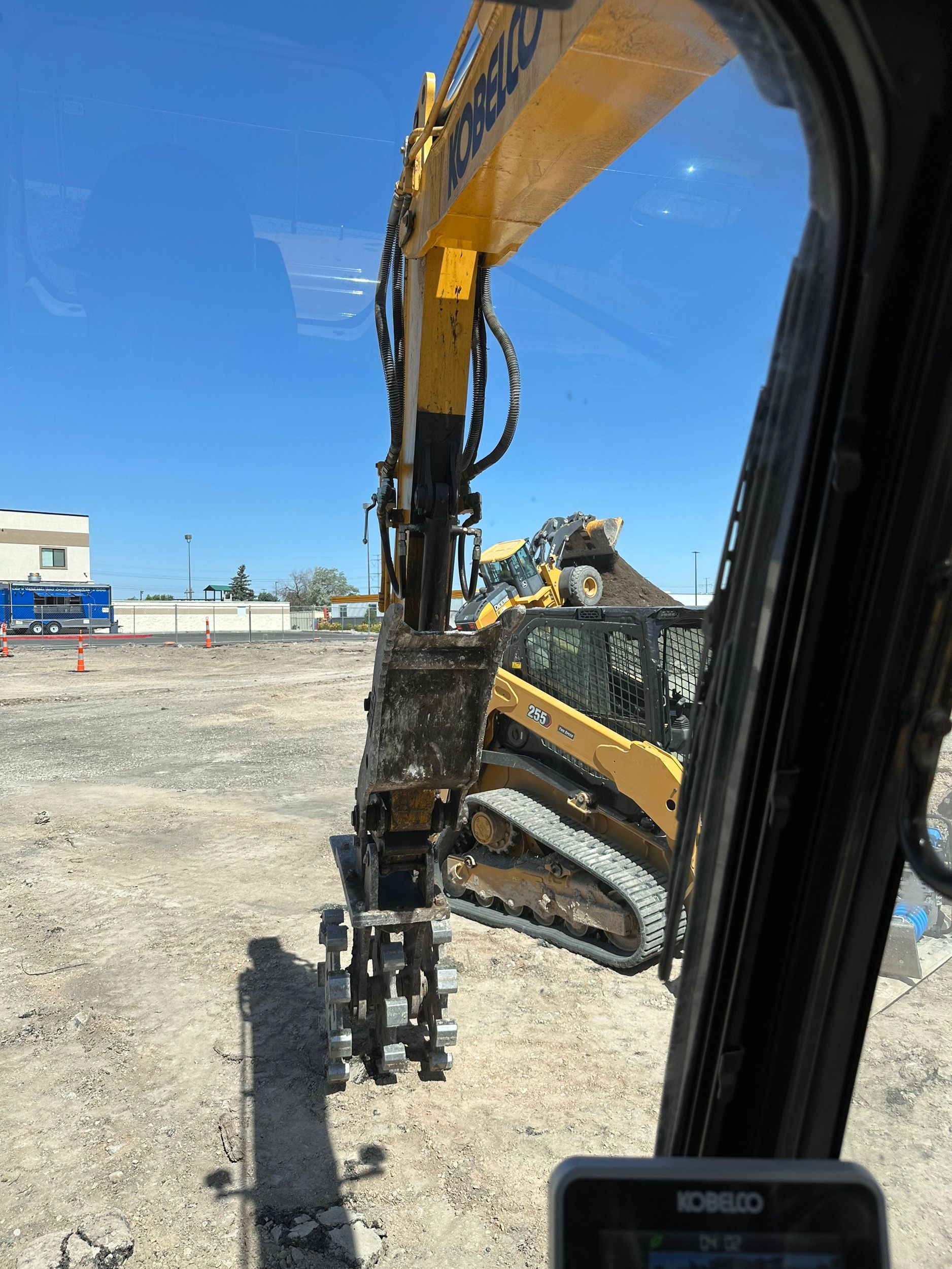 View from inside excavator cab, looking at the boom and a yellow skid steer on a construction site under a clear blue sky.