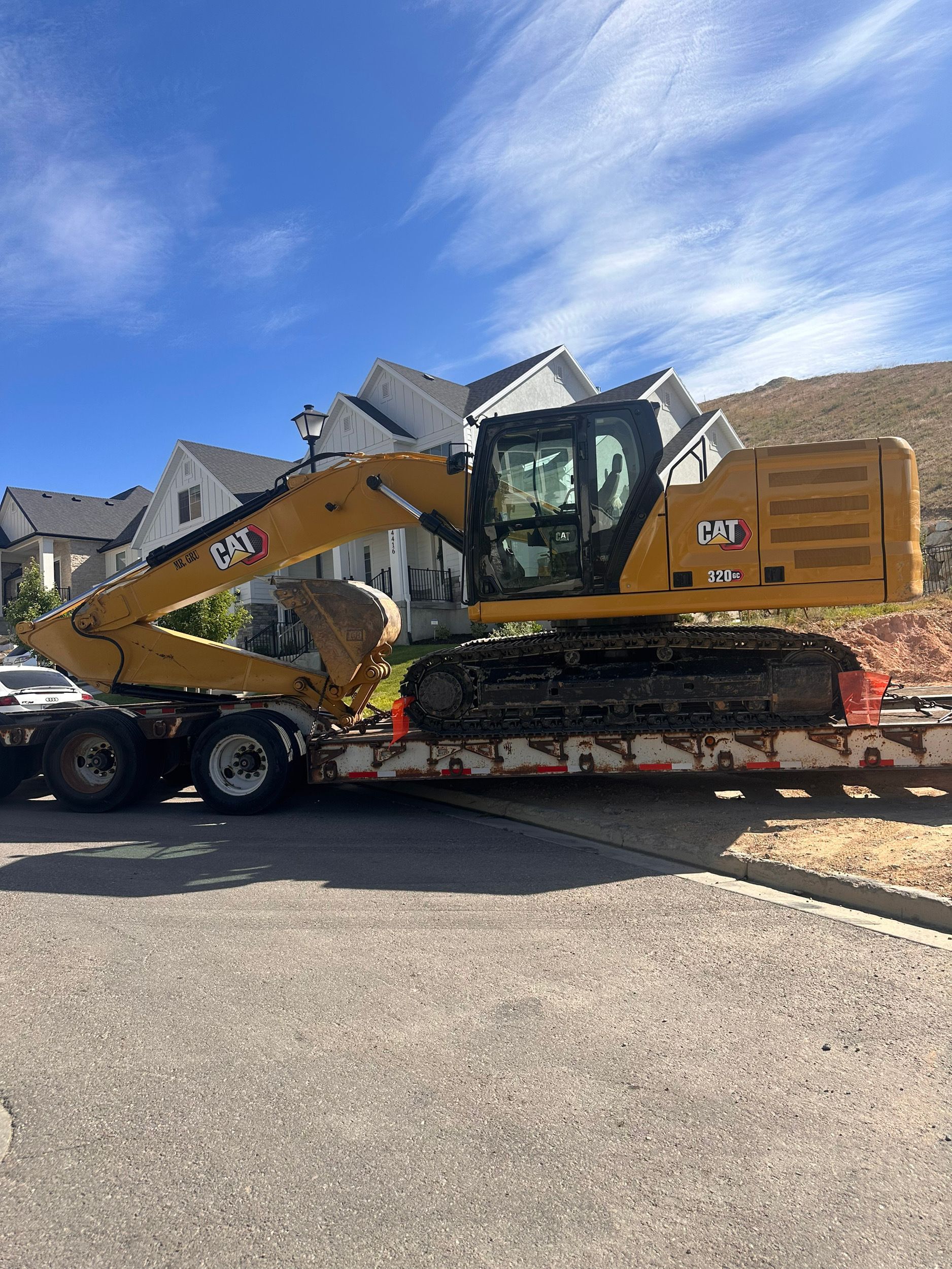 Yellow Caterpillar excavator on a trailer, parked on a paved road with houses and a blue sky in the background.