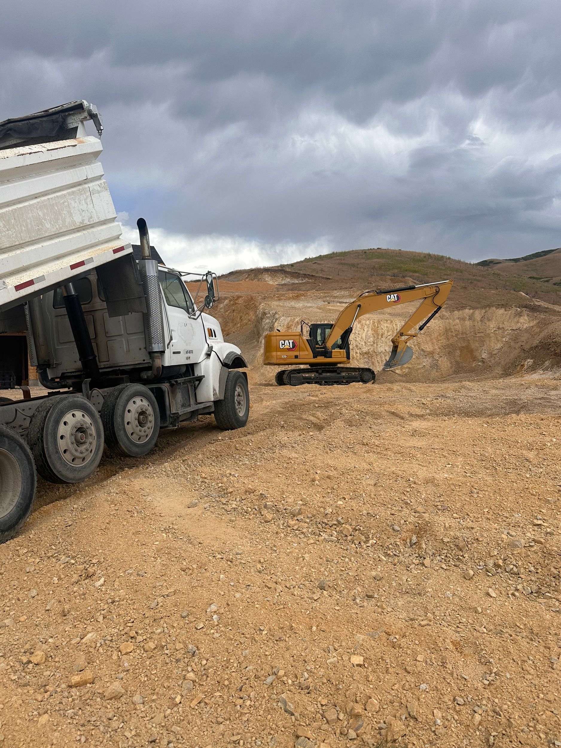 A dump truck unloading gravel near a yellow excavator in a construction zone under a cloudy sky.