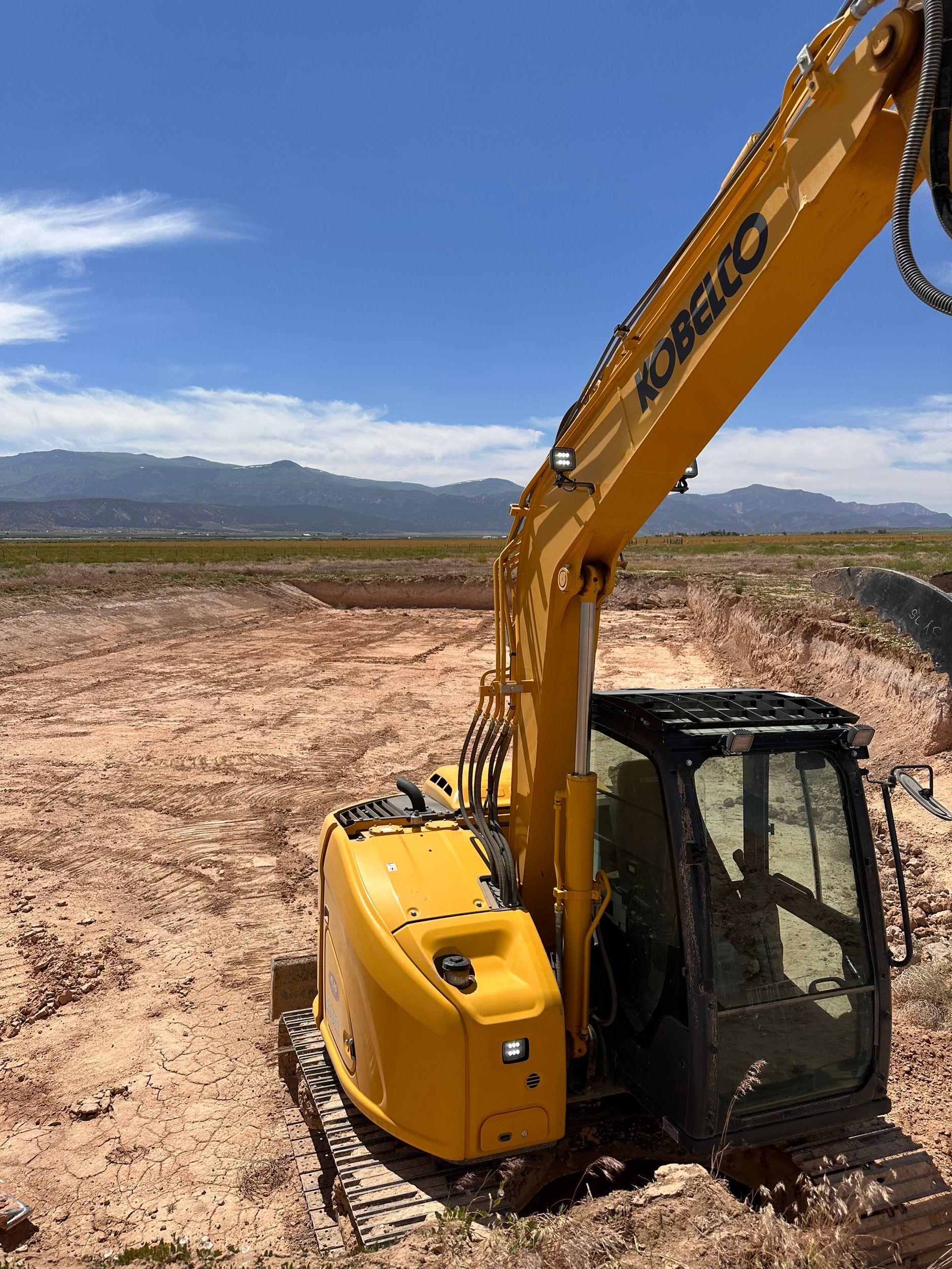 Yellow excavator in a large dirt pit with a mountain range in the background under a blue sky.