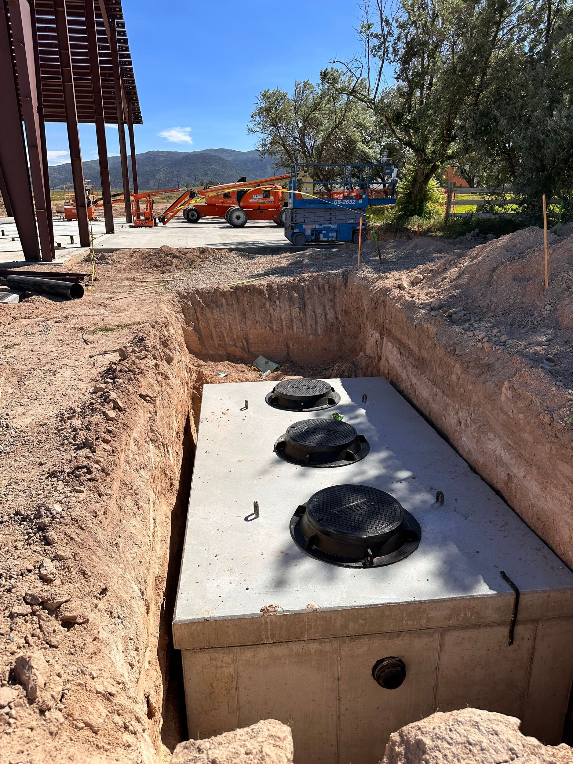 A rectangular concrete septic tank installed in a dirt trench. The tank has three black access covers. Construction equipment is visible in the background.