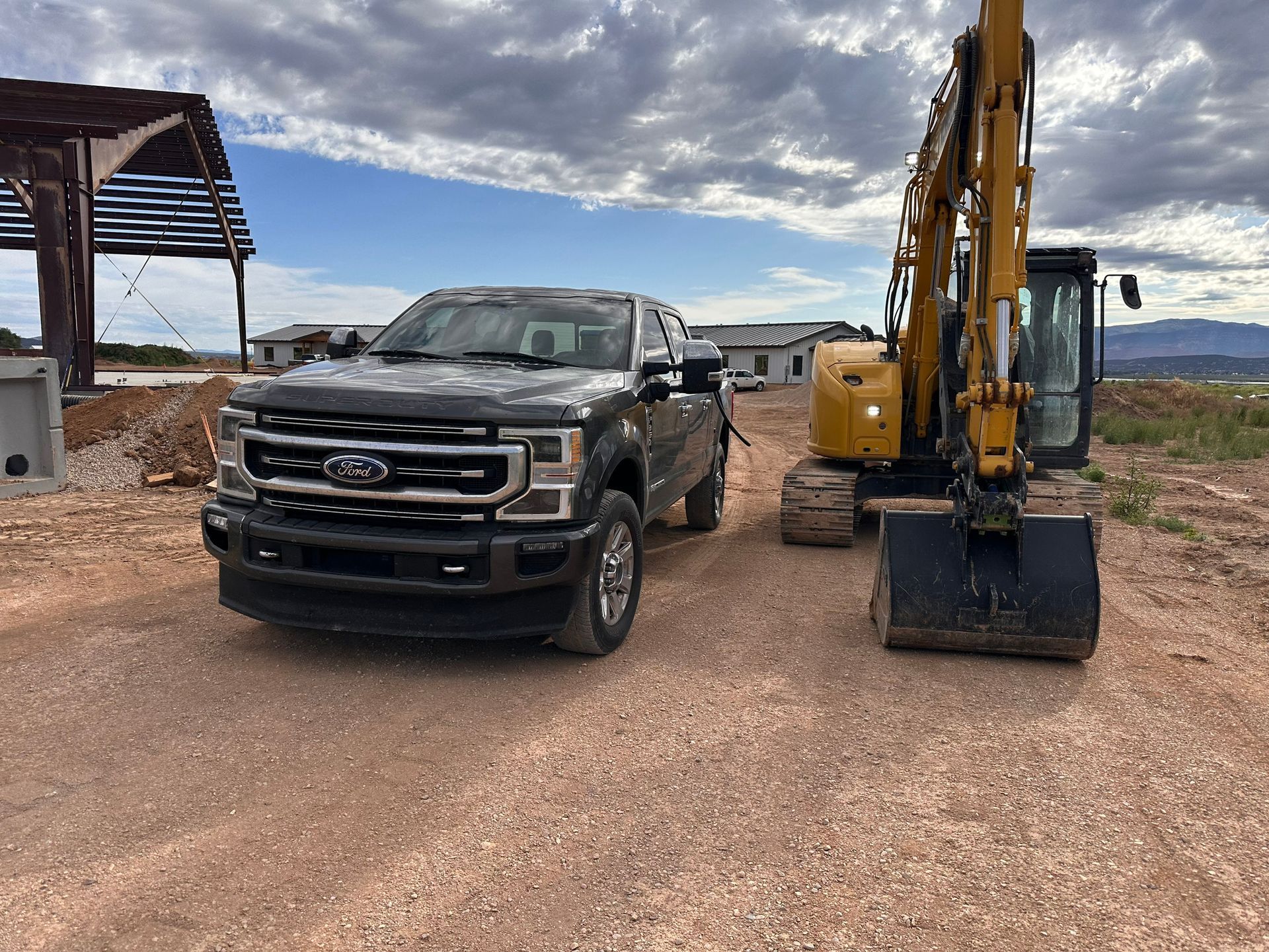 Dark pickup truck parked next to a yellow excavator on a dirt lot, with a cloudy sky in the background.