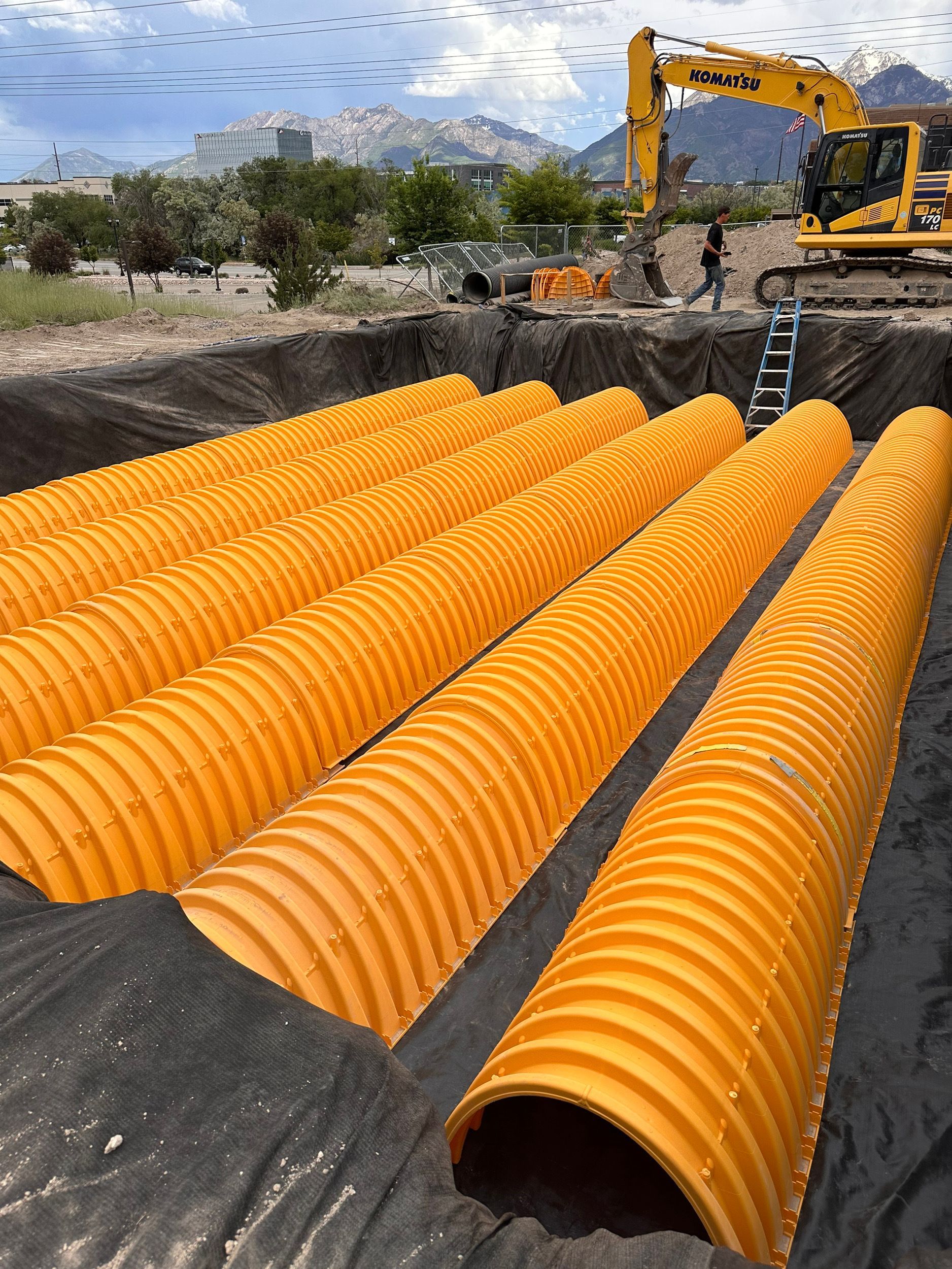 Orange corrugated pipes in a construction site, laid in a deep trench lined with dark fabric. A yellow excavator and mountains are visible in the background.
