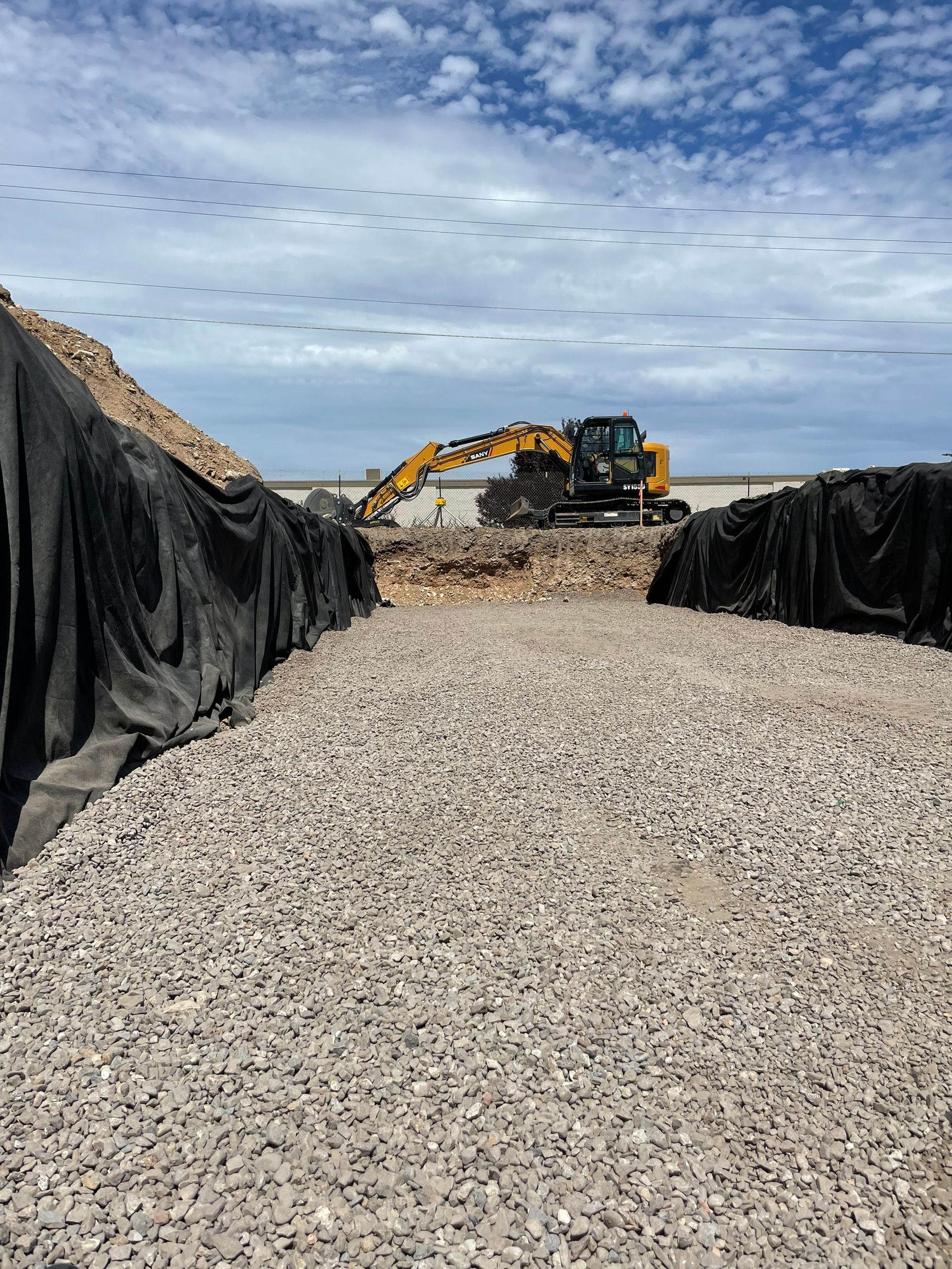 Construction site with a gravel path leading towards an excavator working between two piles of dirt covered in black fabric.