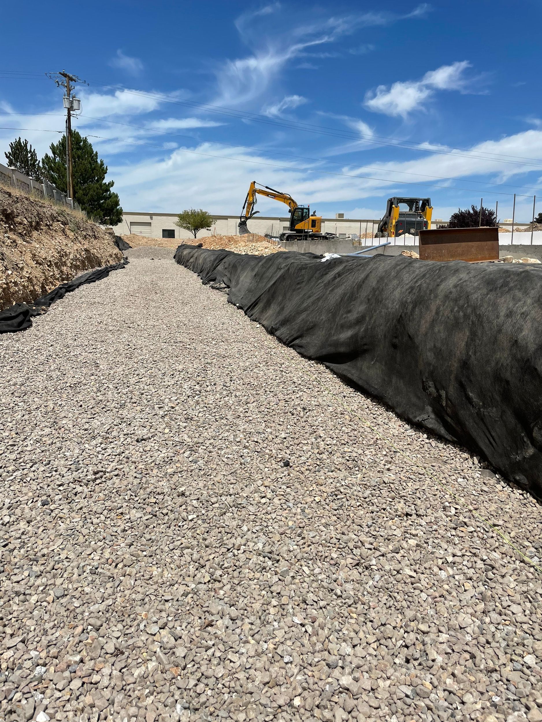 Gravel-lined trench with black lining, construction equipment in the background, under a bright blue sky with scattered clouds.
