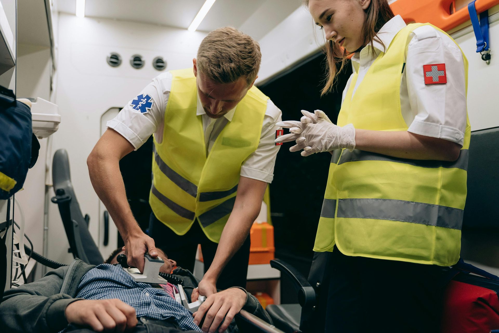 Paramedics treating a patient in an ambulance; man on stretcher, woman assisting, both wearing yellow vests.