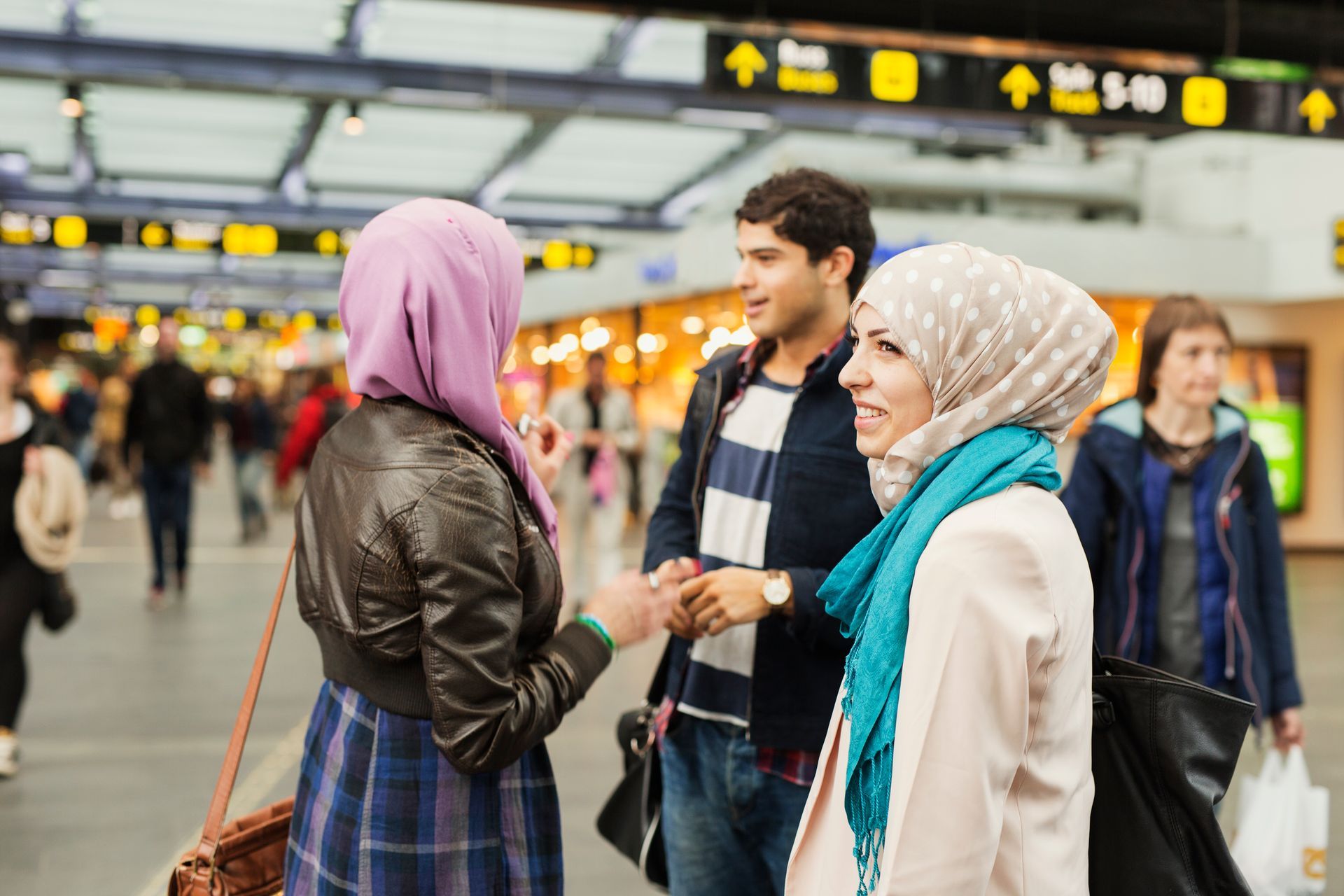 Three young people, two in hijabs, converse in a busy airport terminal.
