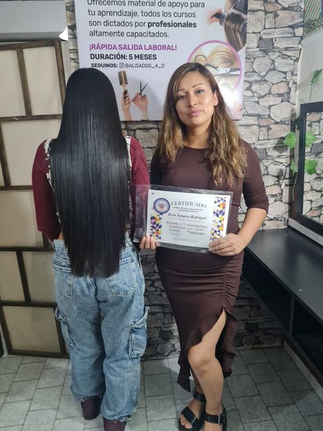 Dos mujeres están de pie en el interior de un edificio; una está de espaldas, la otra sostiene un certificado.