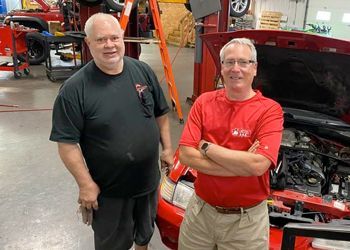 Two men are standing next to each other in front of a red car with the hood open.