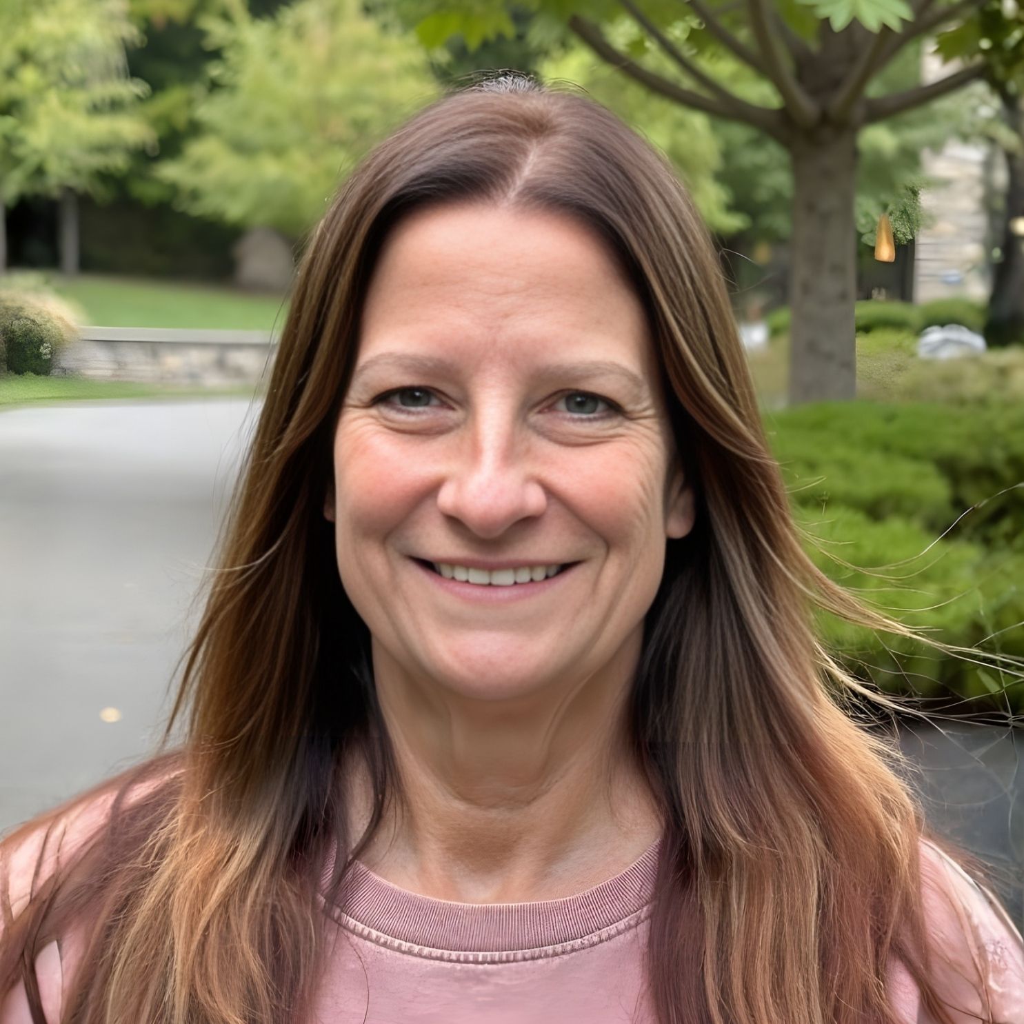 Woman with long brown hair smiles, standing outside, wearing a pink shirt.
