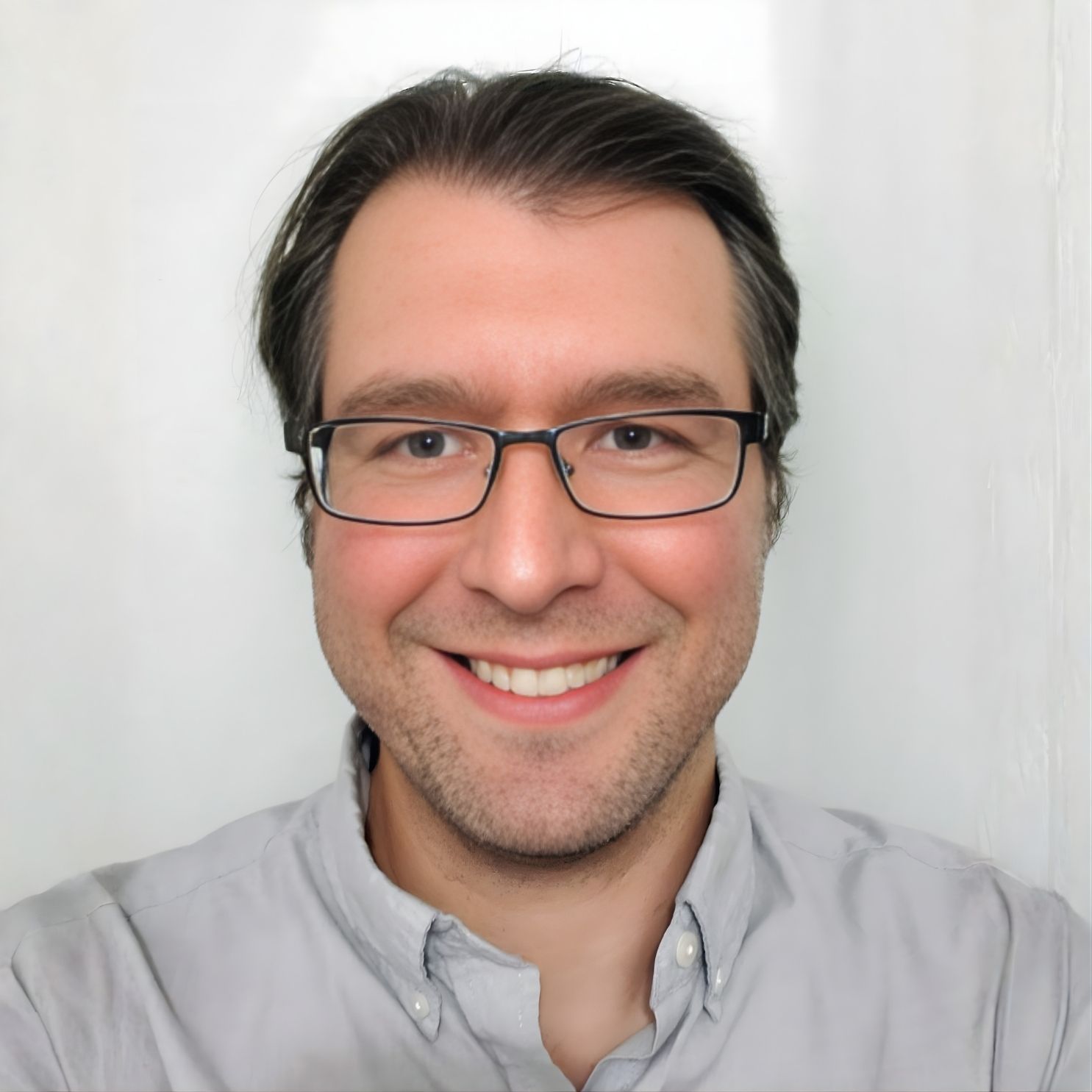 Man with glasses smiling, wearing a gray shirt, against a white backdrop.