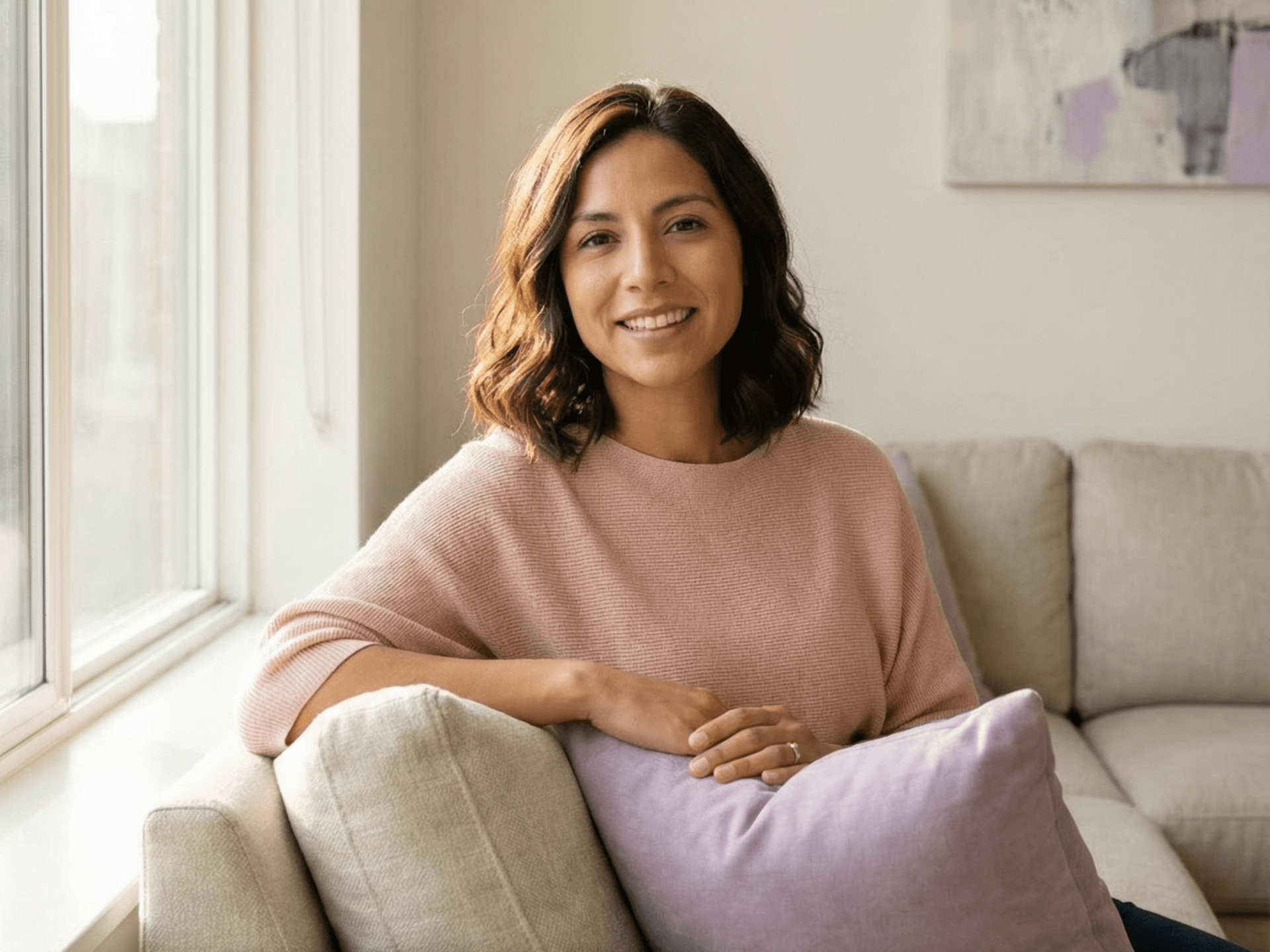 Woman on a couch near a window, smiling. Wearing a pink sweater, holding a purple pillow.