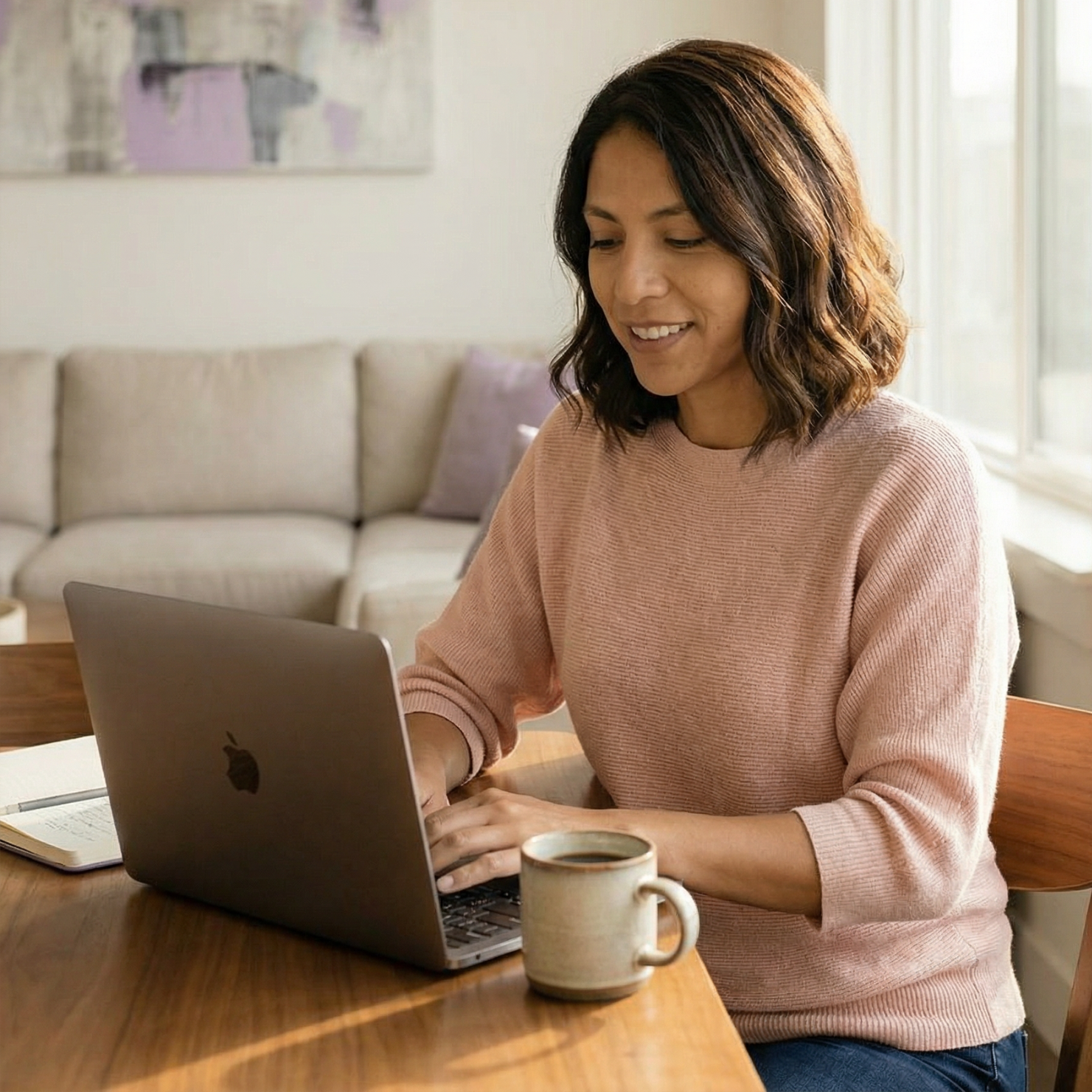 Woman typing on a laptop at a table with a coffee mug; living room setting.
