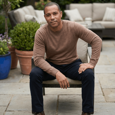 Man seated outdoors, brown sweater, navy pants, looking at camera. Patio setting with plants and seating.