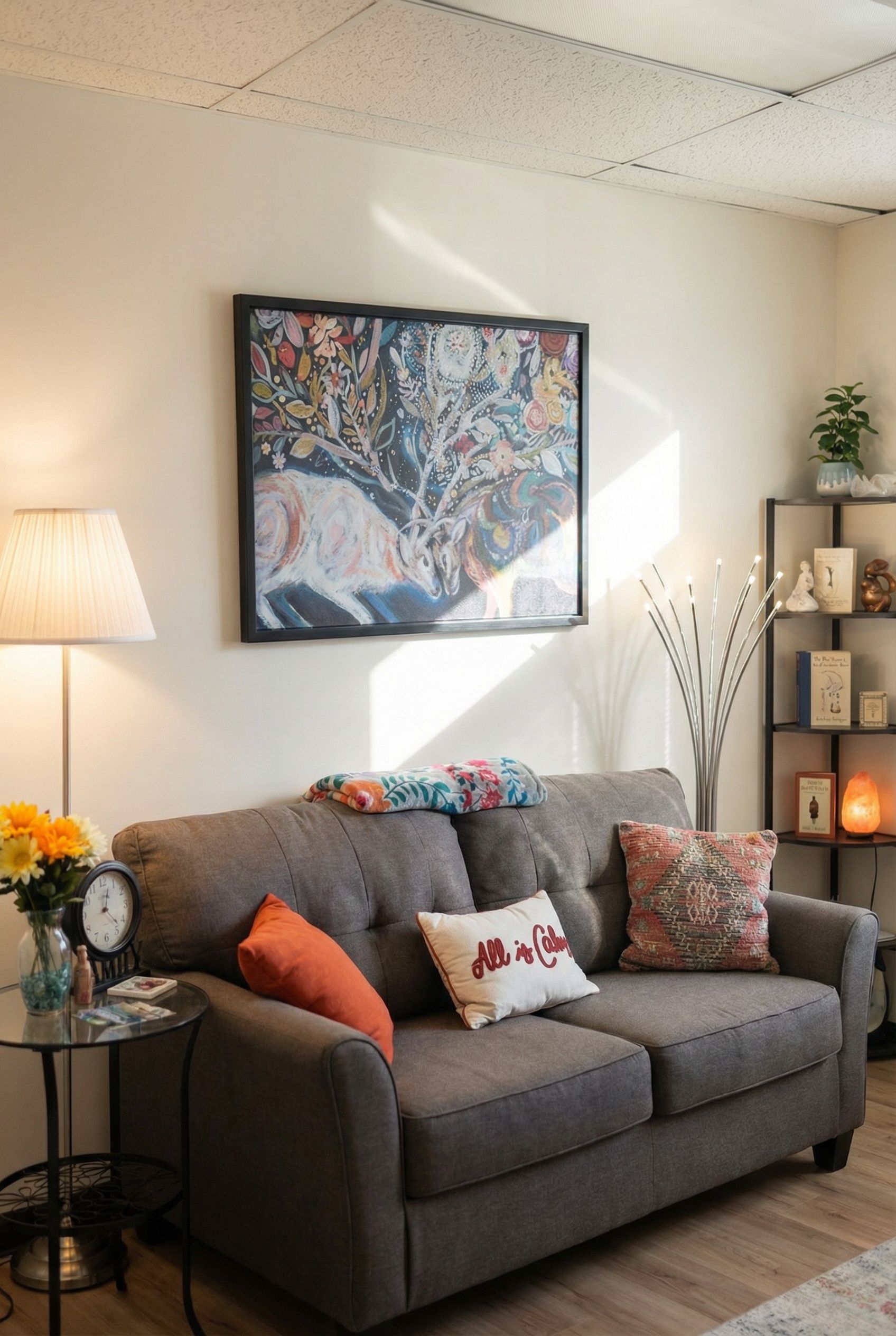 Cozy living room setting, gray sofa with colorful pillows, framed artwork, lamp, side table, and shelving with decor.