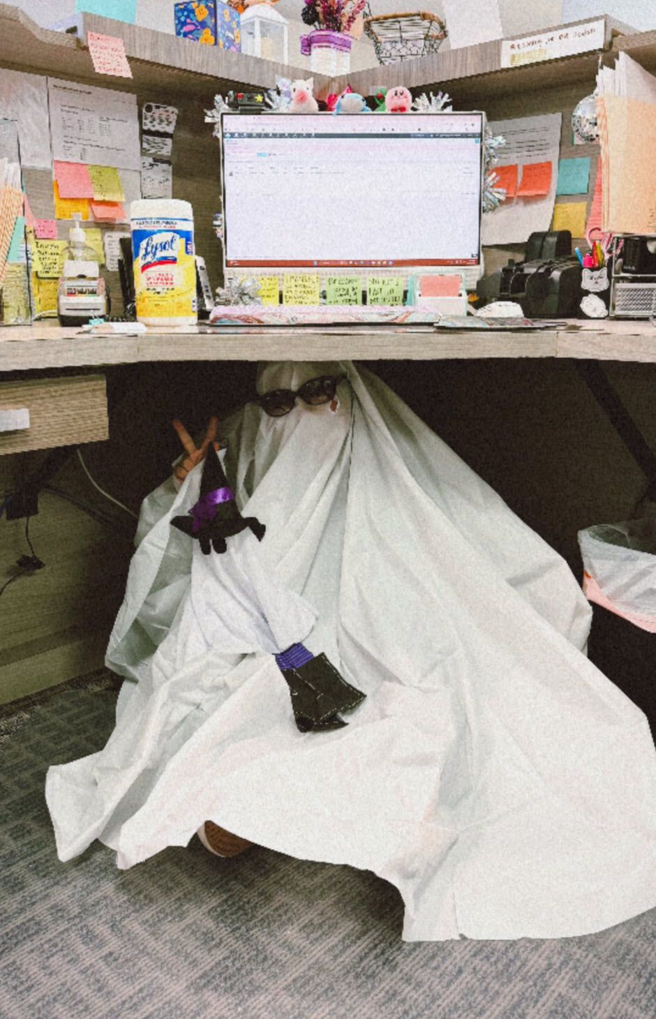 A person in a ghost costume sits under a desk. They hold a small witch hat and glasses. Office setting.