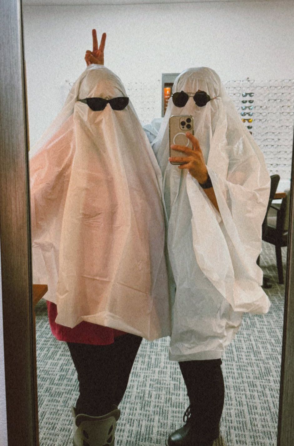 Two people in ghost costumes wearing sunglasses, posing for a mirror selfie indoors. One makes a peace sign.