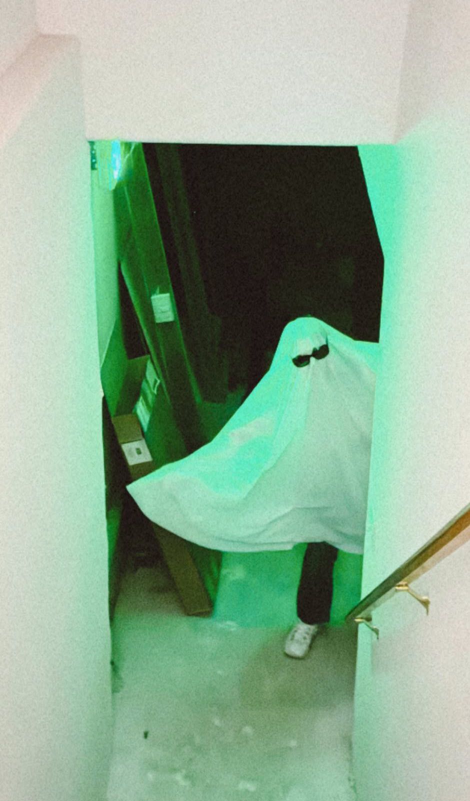 Person in a ghost costume, wearing sunglasses, standing in a doorway with boxes and green lighting.