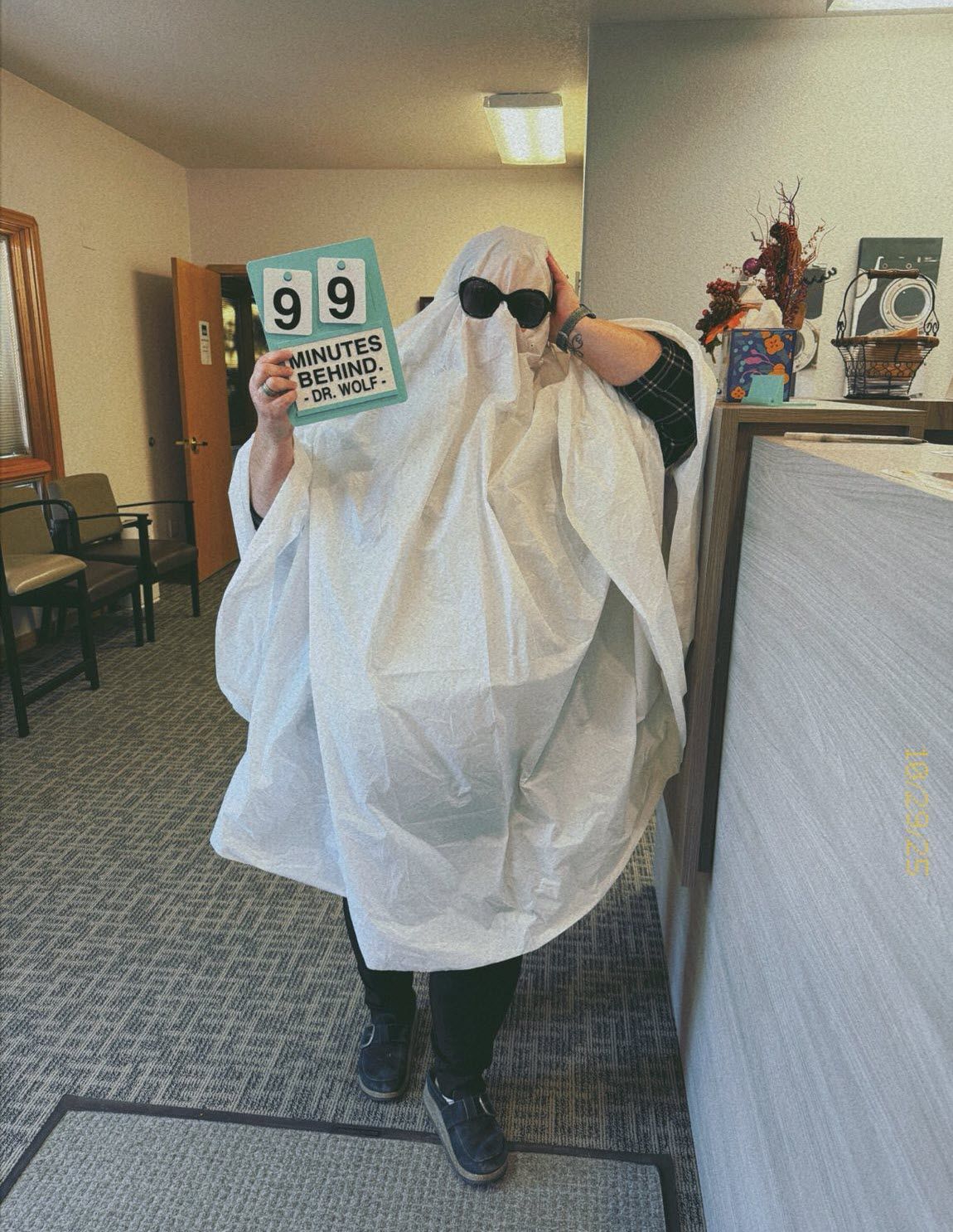 Person in a ghost costume holding a sign with a number 99. They are indoors by a counter.