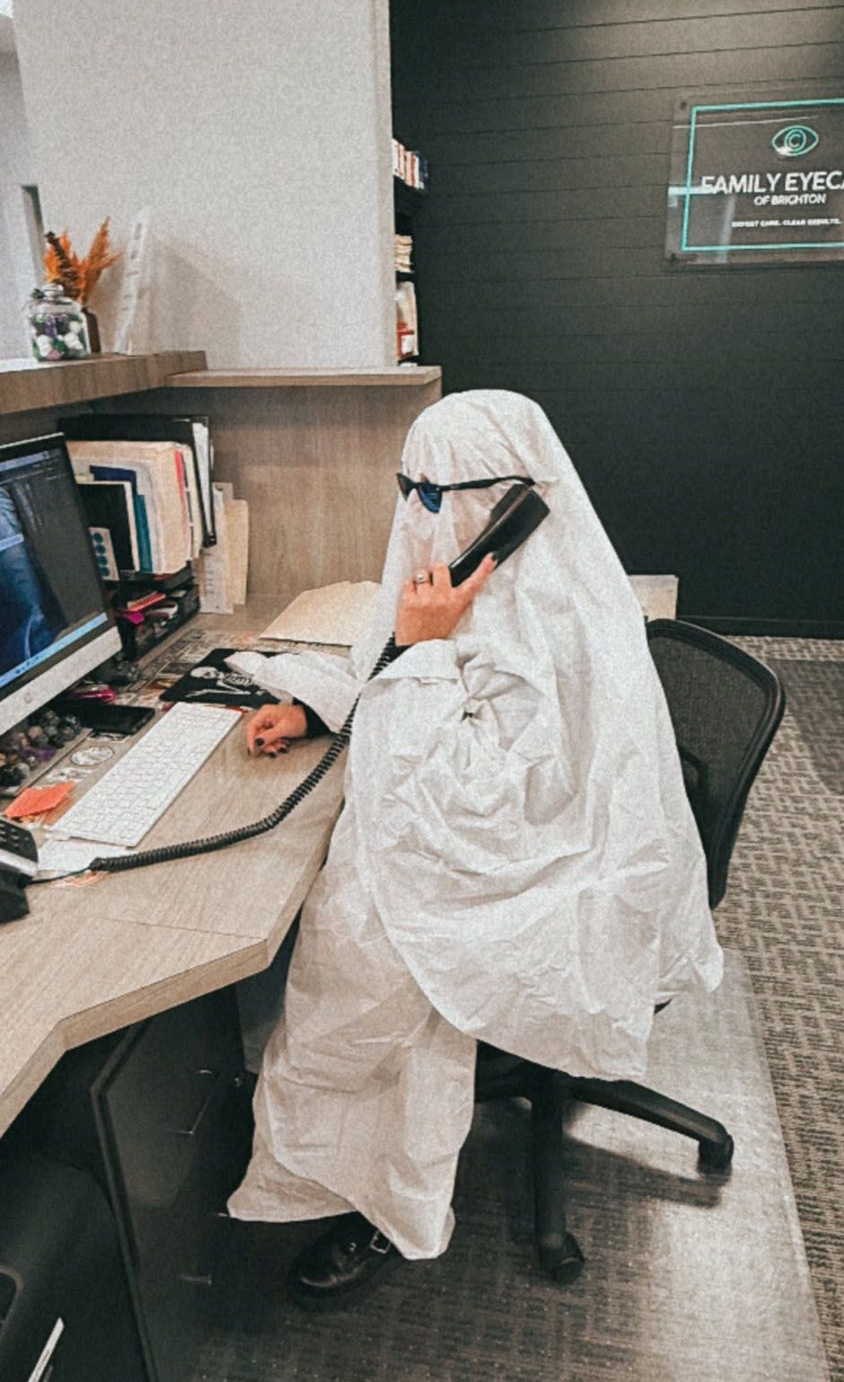 Person in a ghost costume sits at a desk, wearing sunglasses and talking on a phone. Office setting.