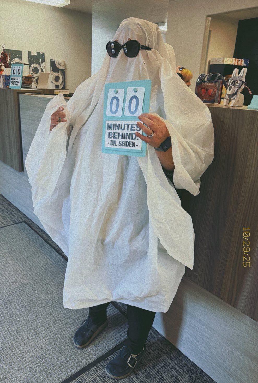 Person in a white ghost costume with sunglasses holding a sign indoors.