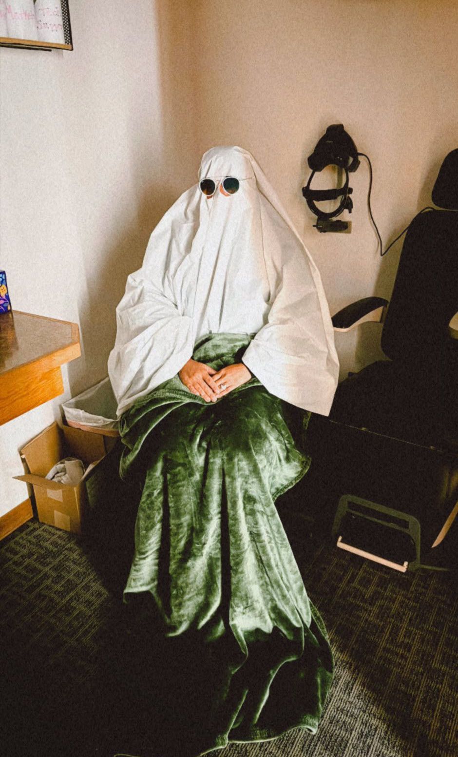 Person in a ghost costume, white sheet and sunglasses, green velvet skirt, seated indoors.