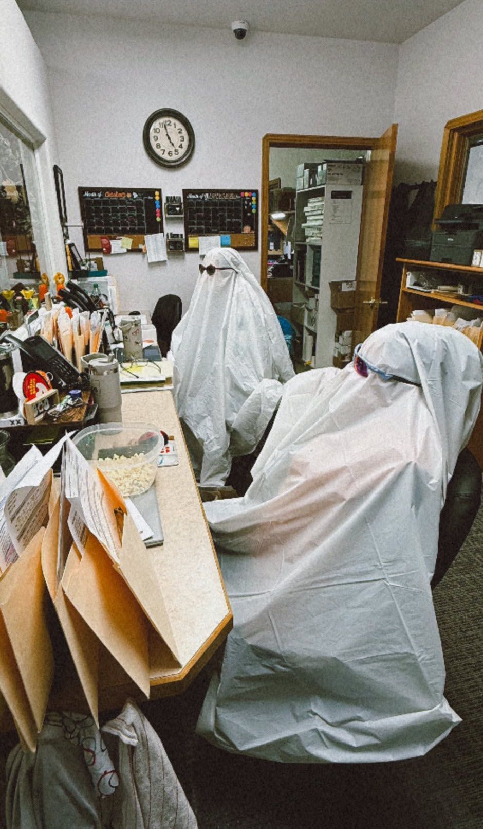Two figures covered in white sheets in an office, desk with papers, clock on wall.