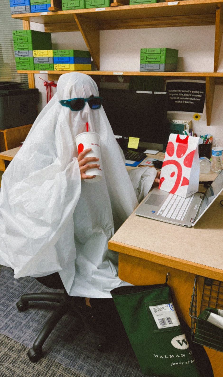A person dressed as a ghost, wearing sunglasses, drinks a beverage from Chick-fil-A while at a desk with a laptop.
