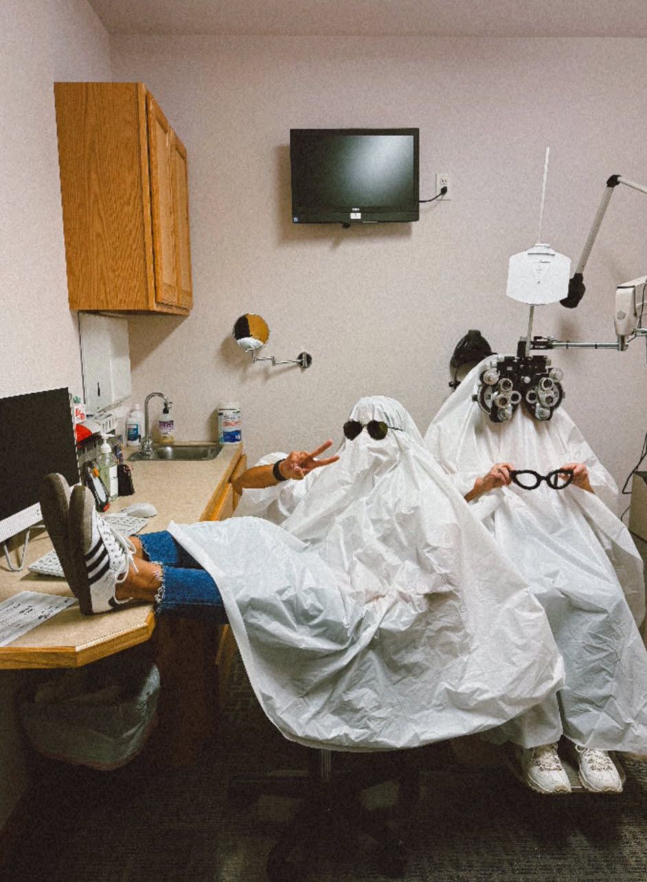 Two people in ghost costumes at an eye doctor's office, one with feet on a desk, the other with eye exam glasses.