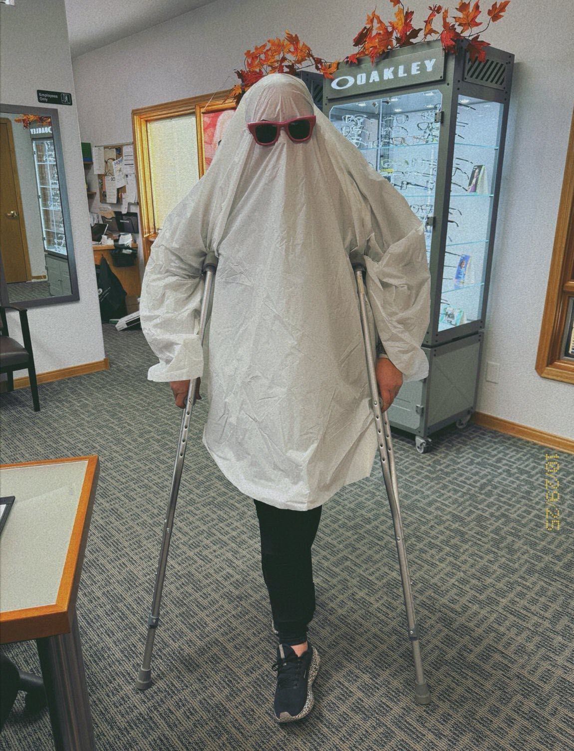 Person in a white ghost costume with sunglasses using crutches inside an eyeglass store.