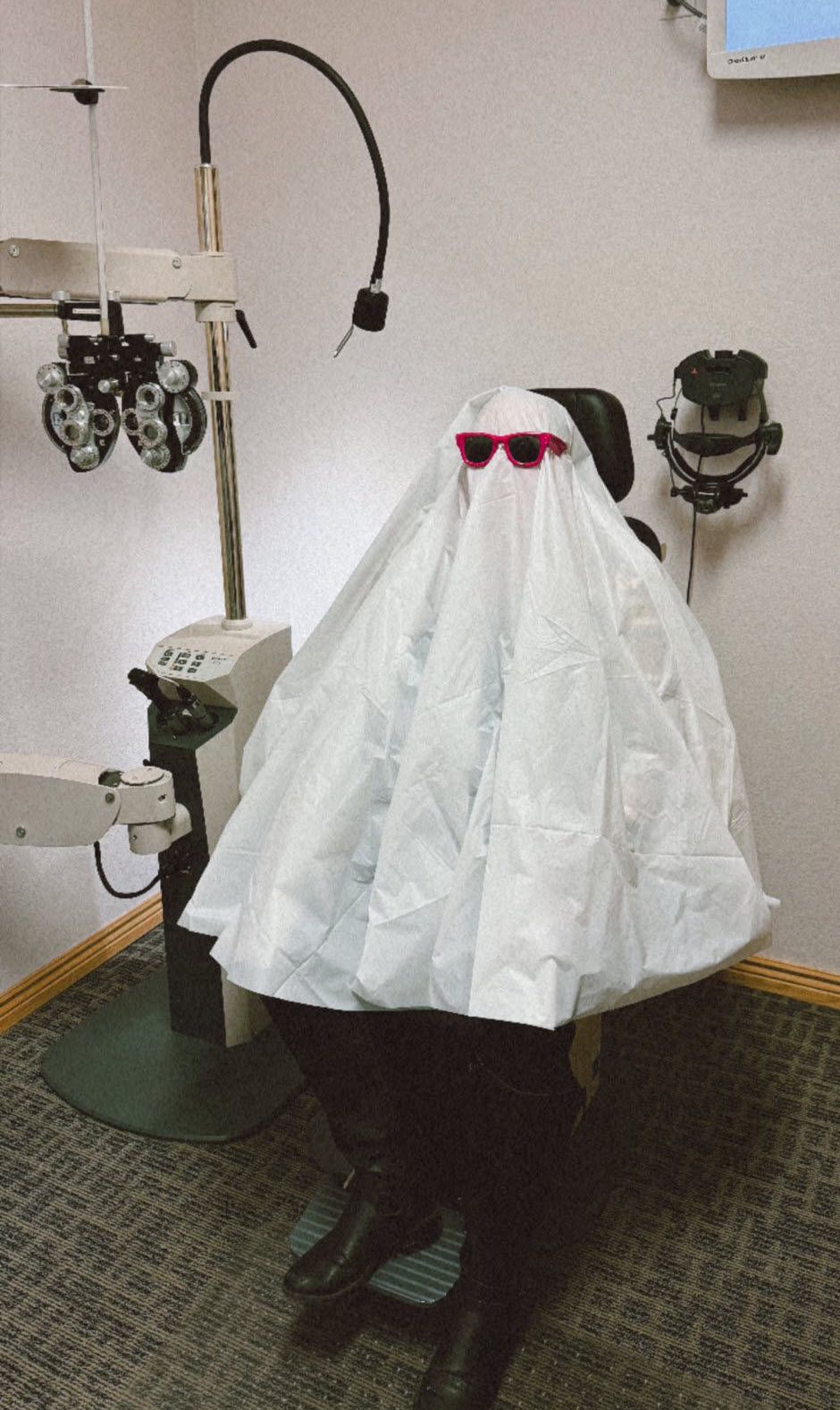 Person dressed as a ghost in an eye doctor's chair wearing red sunglasses.