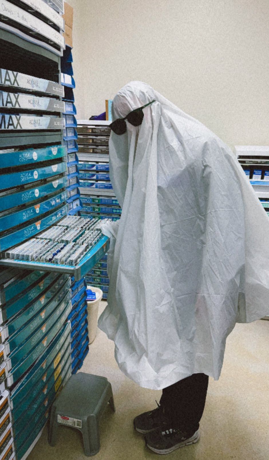 Person in a ghost costume wearing sunglasses, examining items on shelves in a supply room.