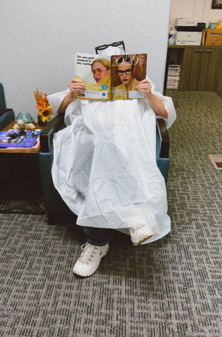 Person in a medical gown sits in a chair, reading a magazine. Office setting.