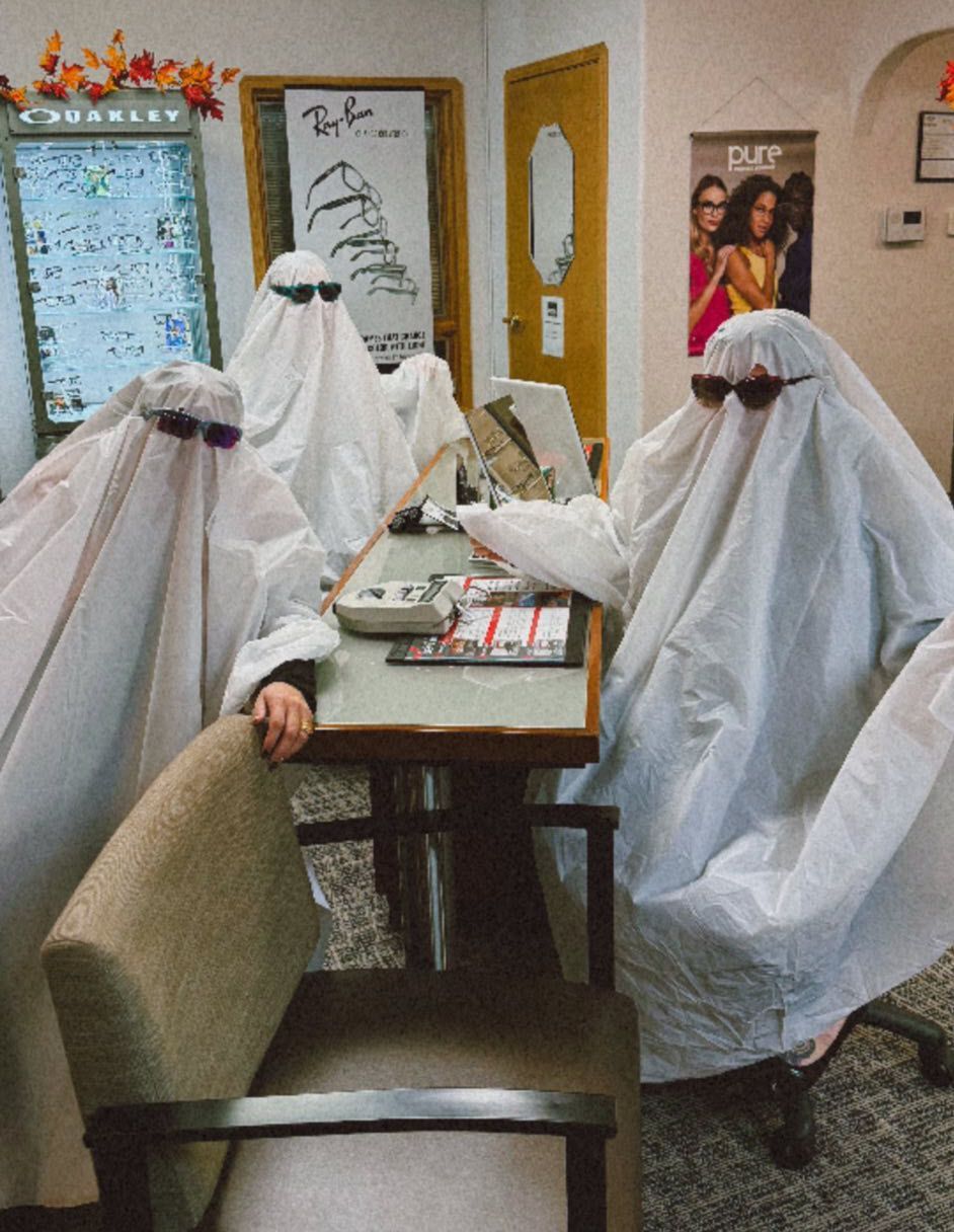 Three people in ghost costumes wearing sunglasses at a desk in an optical shop.