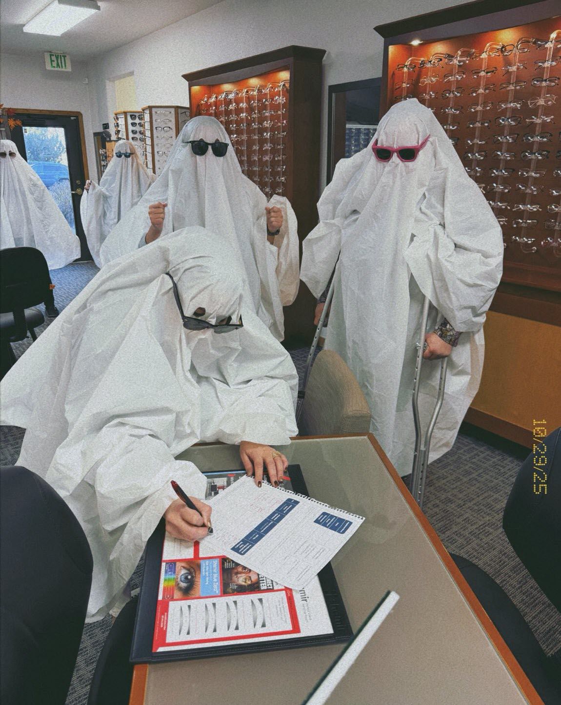 People in ghost costumes, wearing sunglasses, at an optometrist's office. One ghost signs paperwork.
