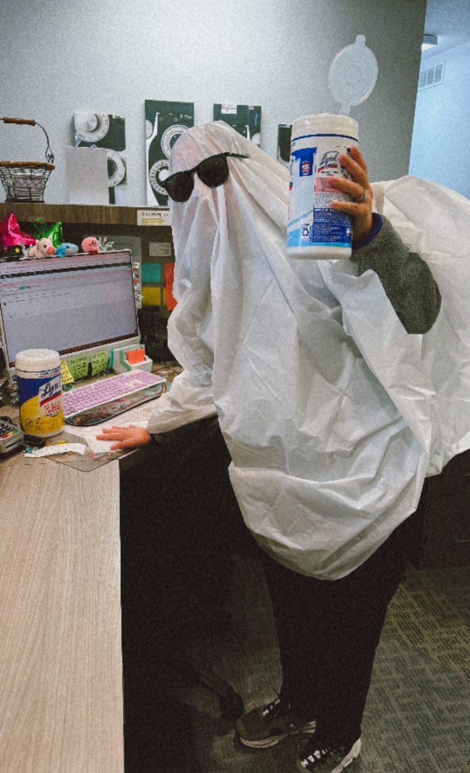 Person in ghost costume, wearing sunglasses, holding sanitizing wipes by a desk.