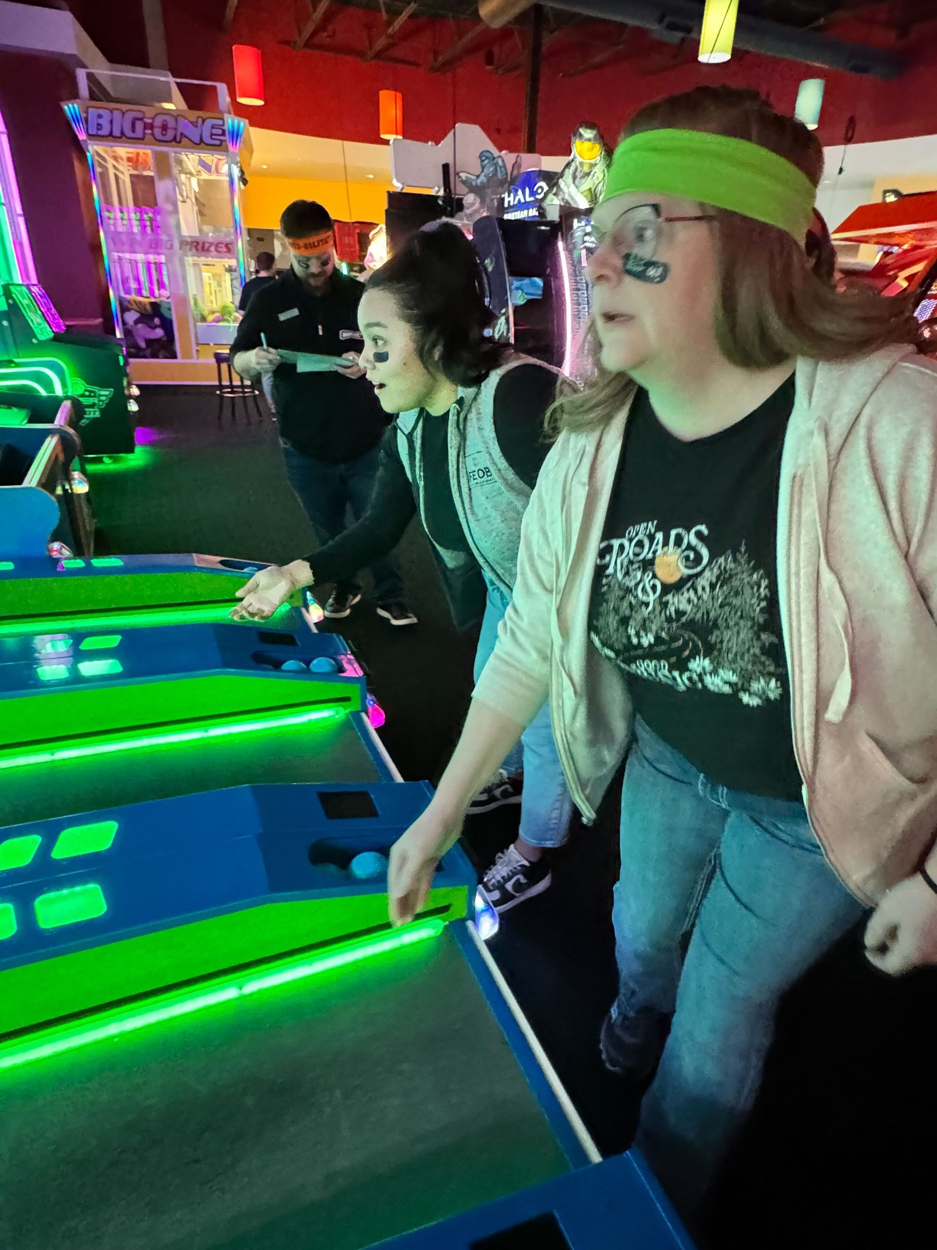 Three people playing arcade skee-ball, two women in foreground, all wearing headbands, lit by neon green lights.