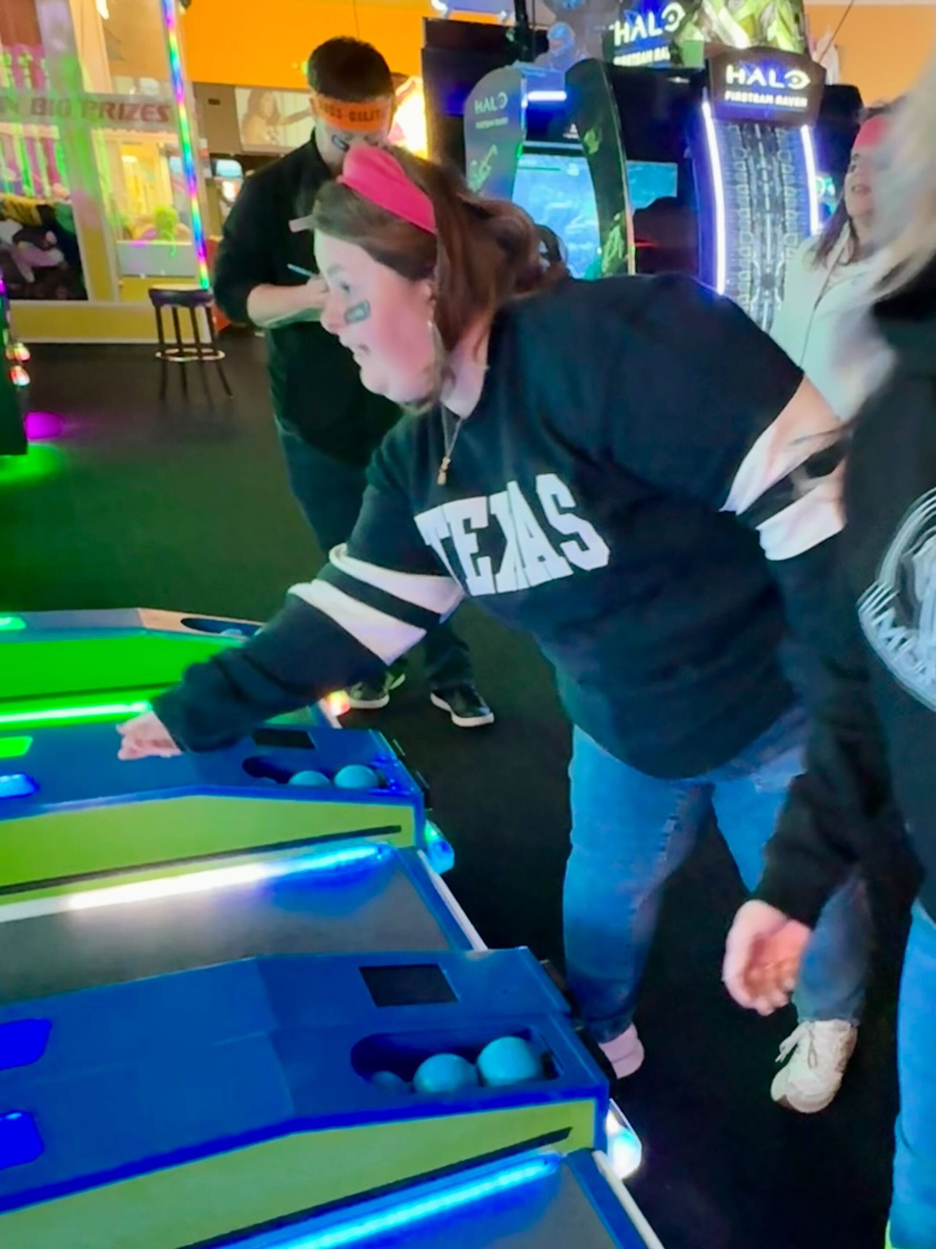 Woman in Texas shirt playing an arcade game, reaching out, with another person nearby.
