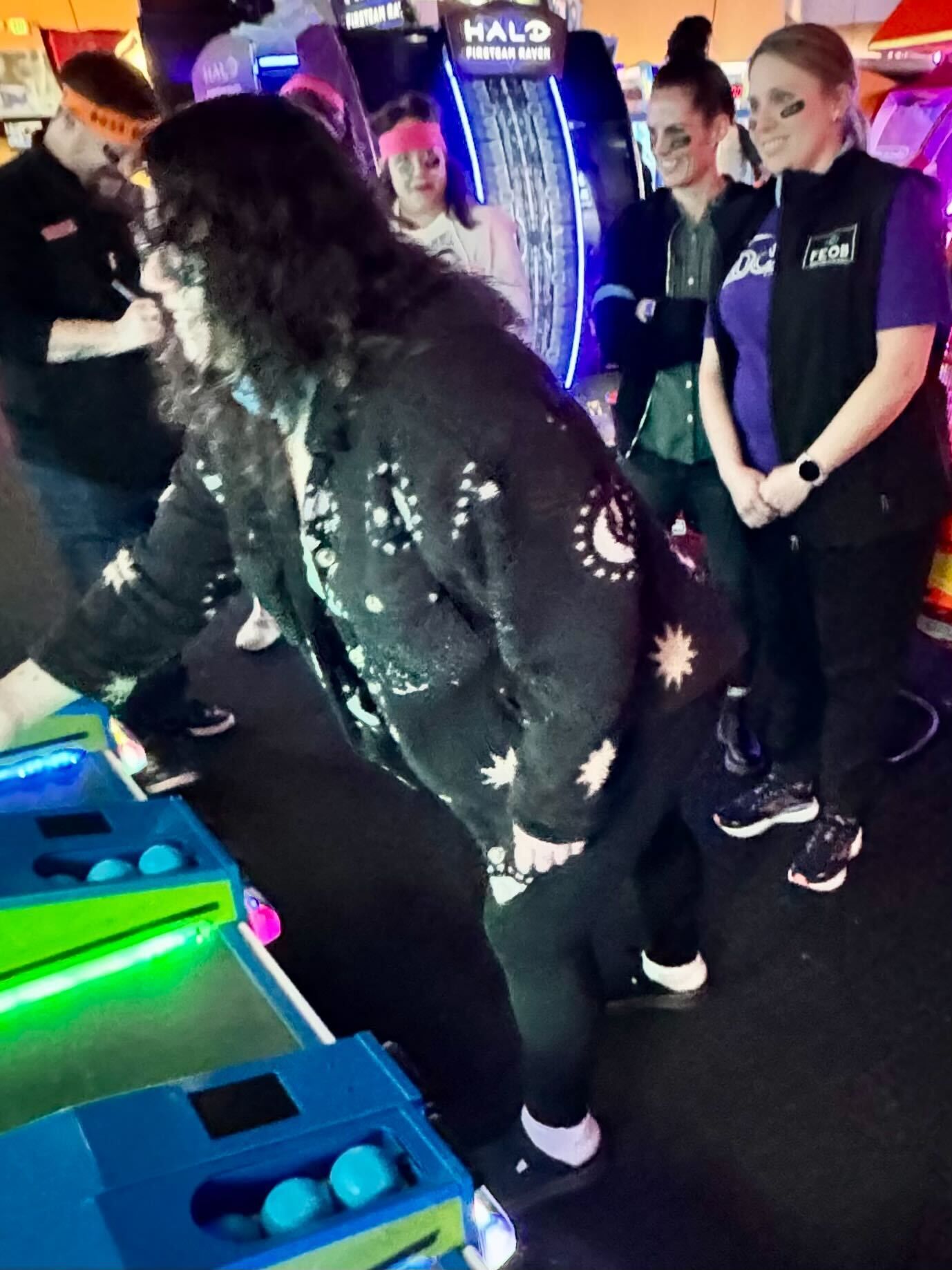A woman in a patterned sweater plays an arcade game. Others watch in a game room.